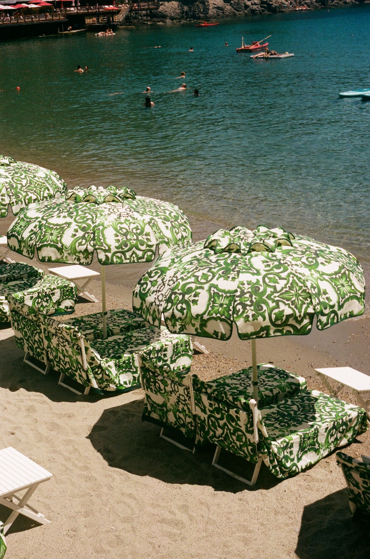 Green and white patterned beach umbrellas with matching lounge chairs on a sandy Mediterranean beach with turquoise water.