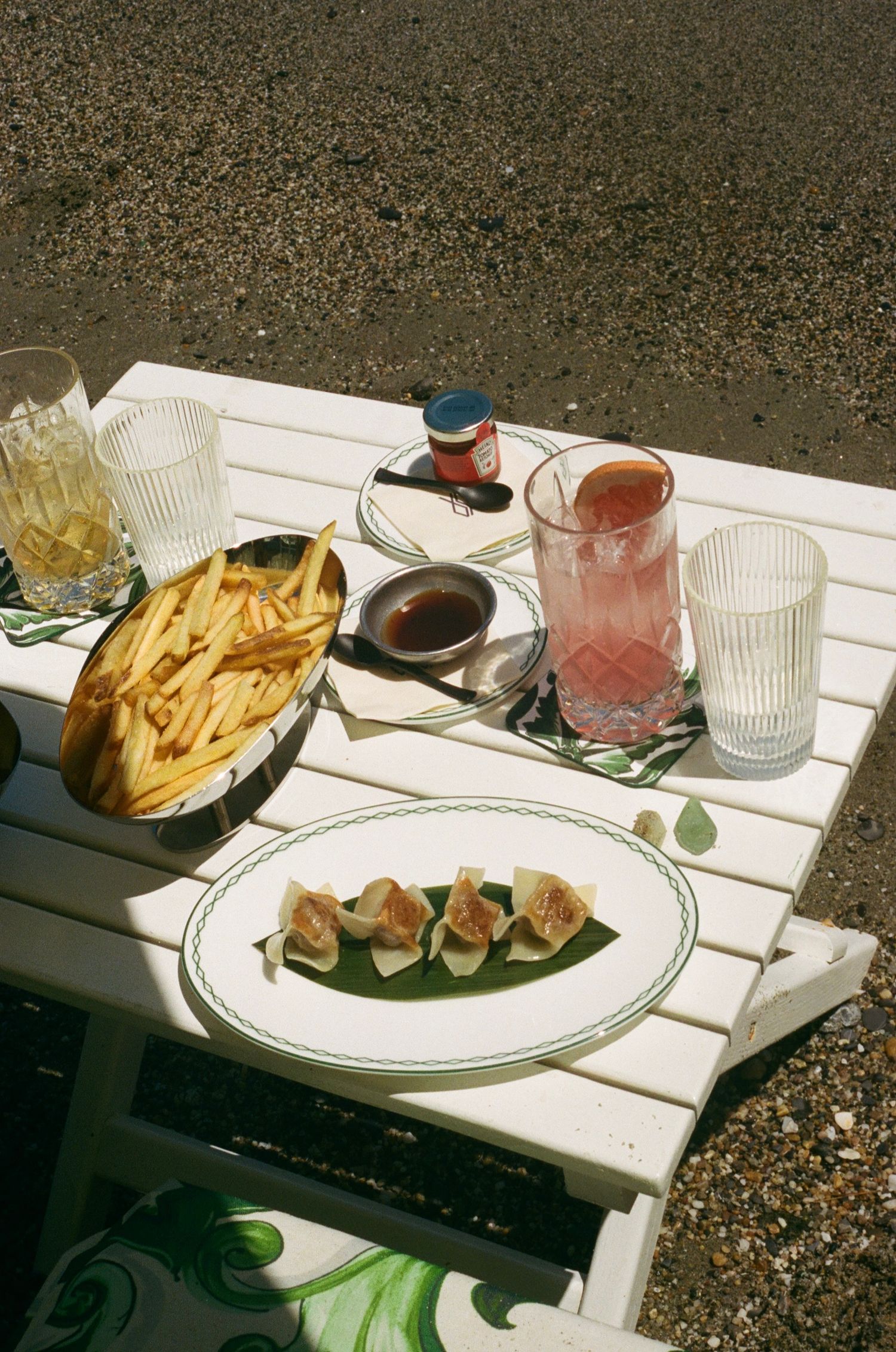 Outdoor white table with dumplings, french fries, dipping sauce, pink cocktail, and water glass on gravel.