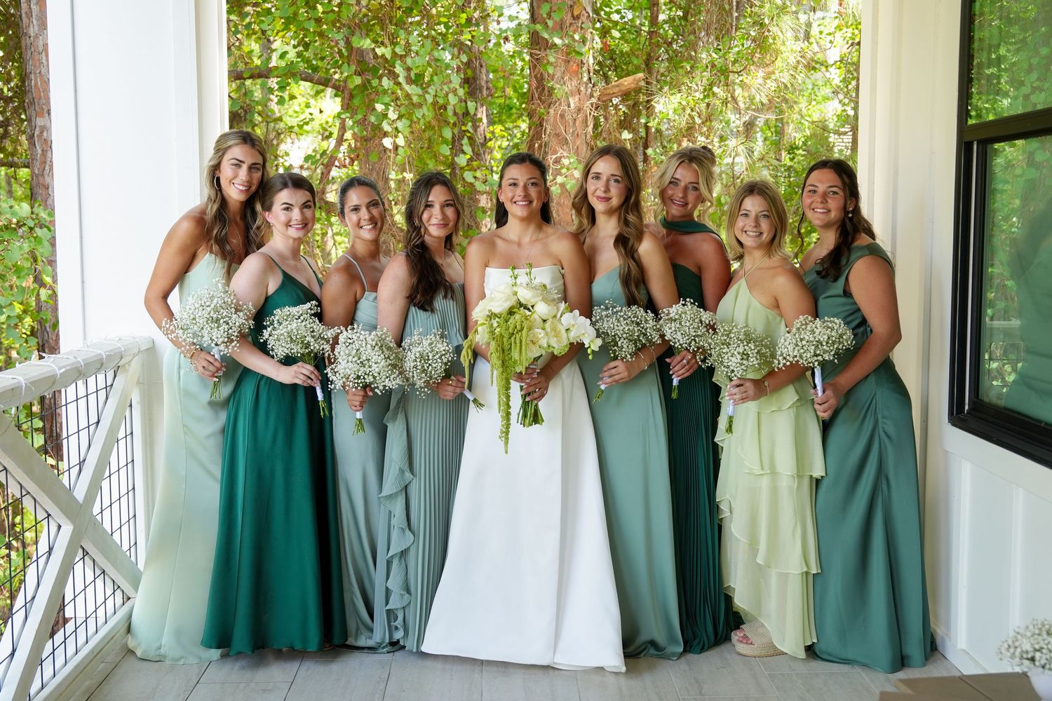 Bride in white gown with bridesmaids in mismatched green dresses holding white floral bouquets on a porch.