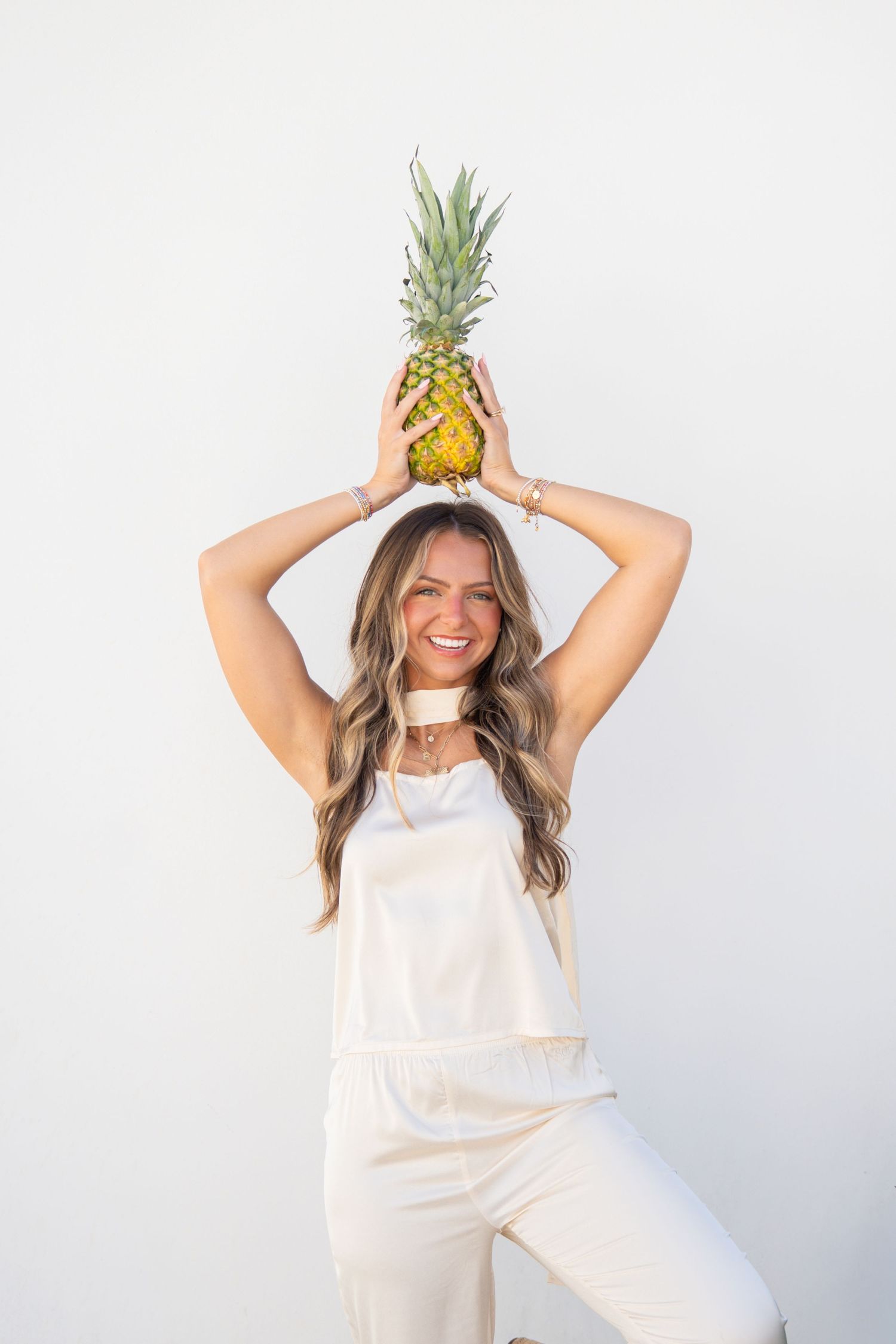 Smiling woman in white overalls holding a pineapple above her head against a white background.