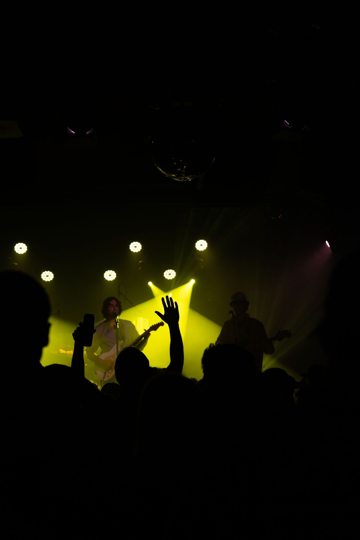Concert crowd silhouettes against yellow stage lighting raise their hands in celebration during a live performance.