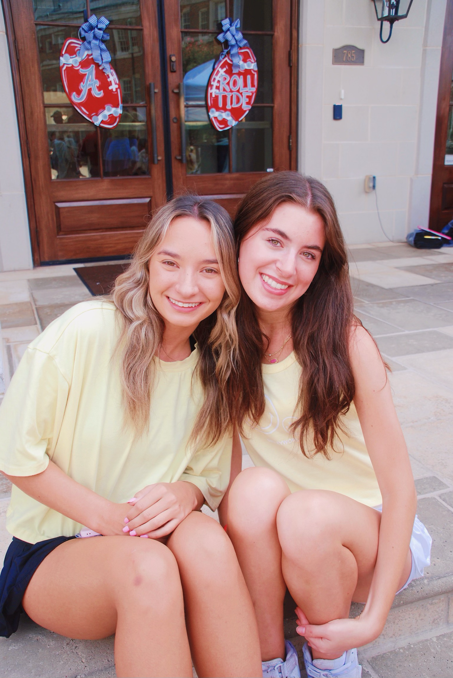 Two friends in matching yellow shirts sit together on front steps with heart-shaped decorations visible on the door behind them.