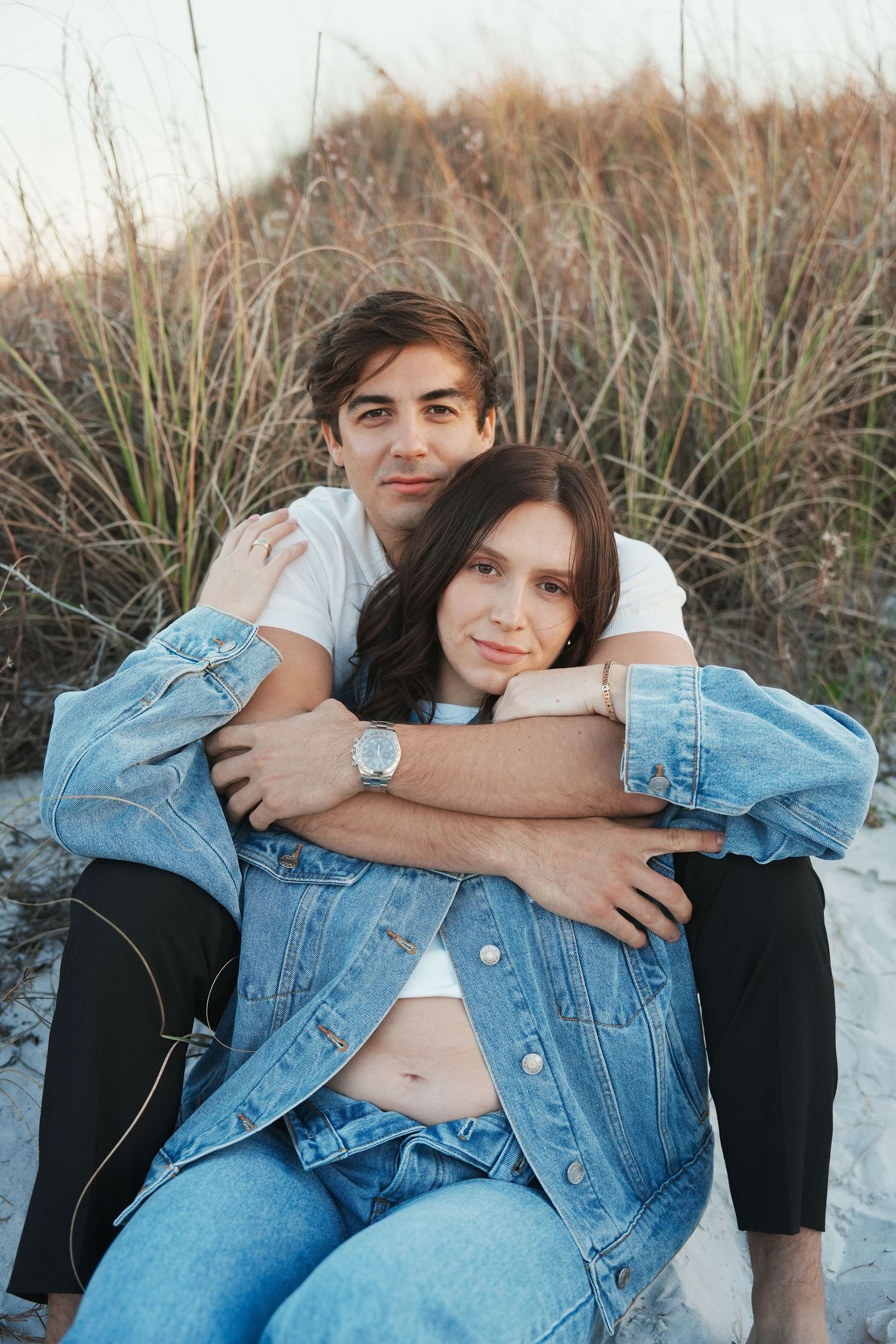 A couple in denim and white sits together on a sandy beach at sunset in a series of intimate poses.