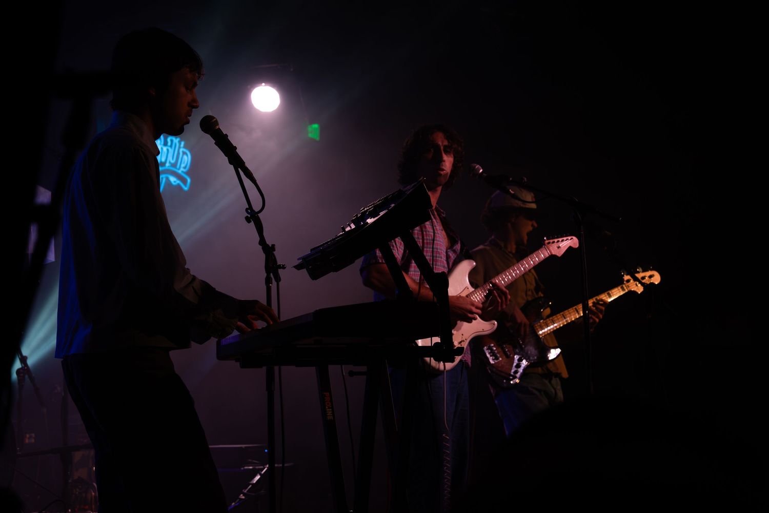 Silhouetted musicians perform on a dimly lit stage with dramatic blue lighting and a bright moon-like backdrop.