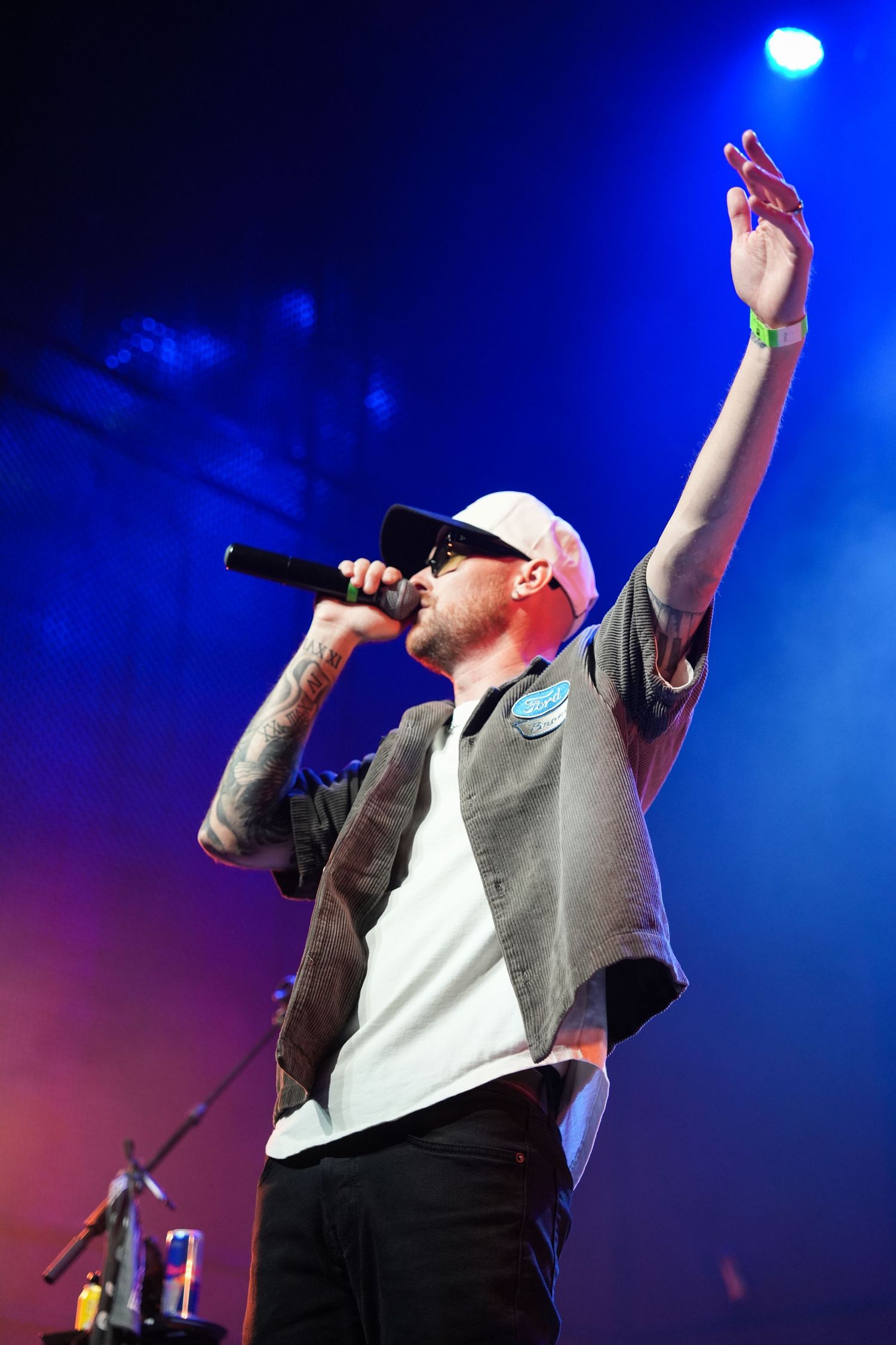 Performer in white baseball cap and vest singing into microphone with raised arm on stage under dramatic blue lighting.