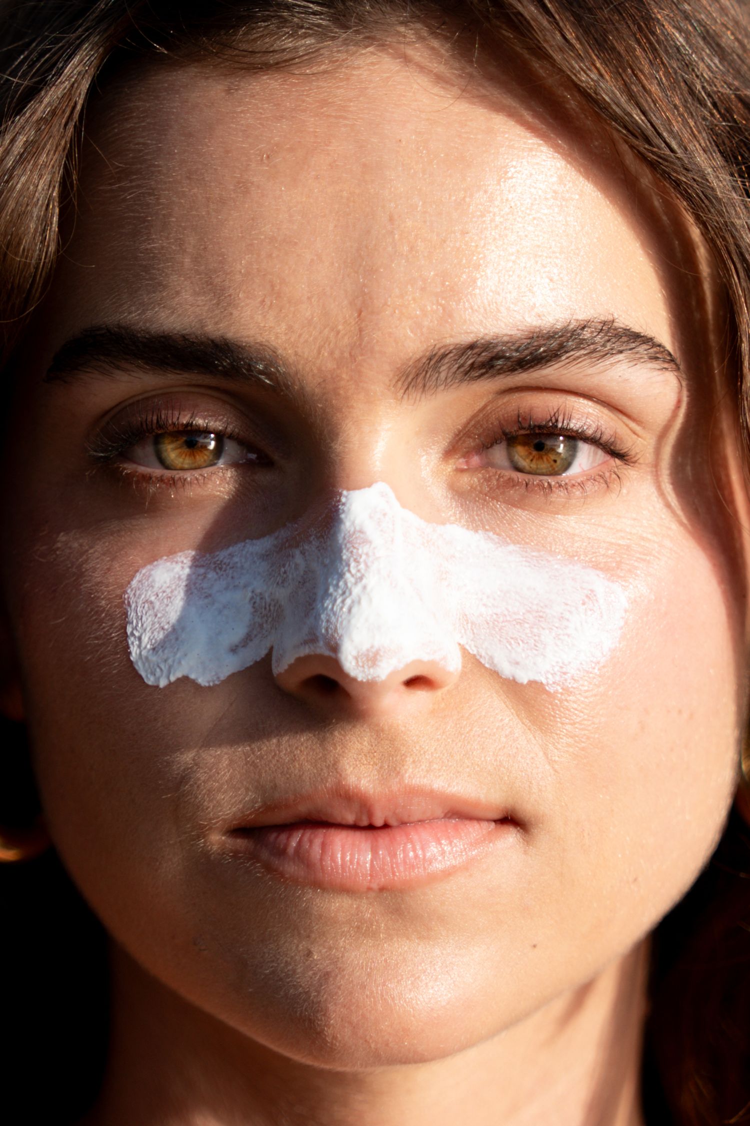 Close-up portrait of a woman with white sunscreen applied across her nose and cheeks in bright sunlight.