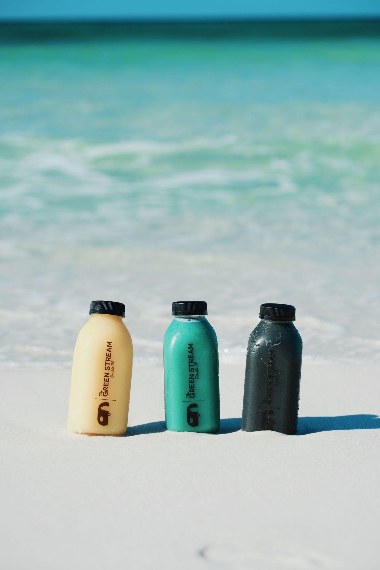 Three colorful cosmetic containers displayed on white sand with turquoise ocean water background.