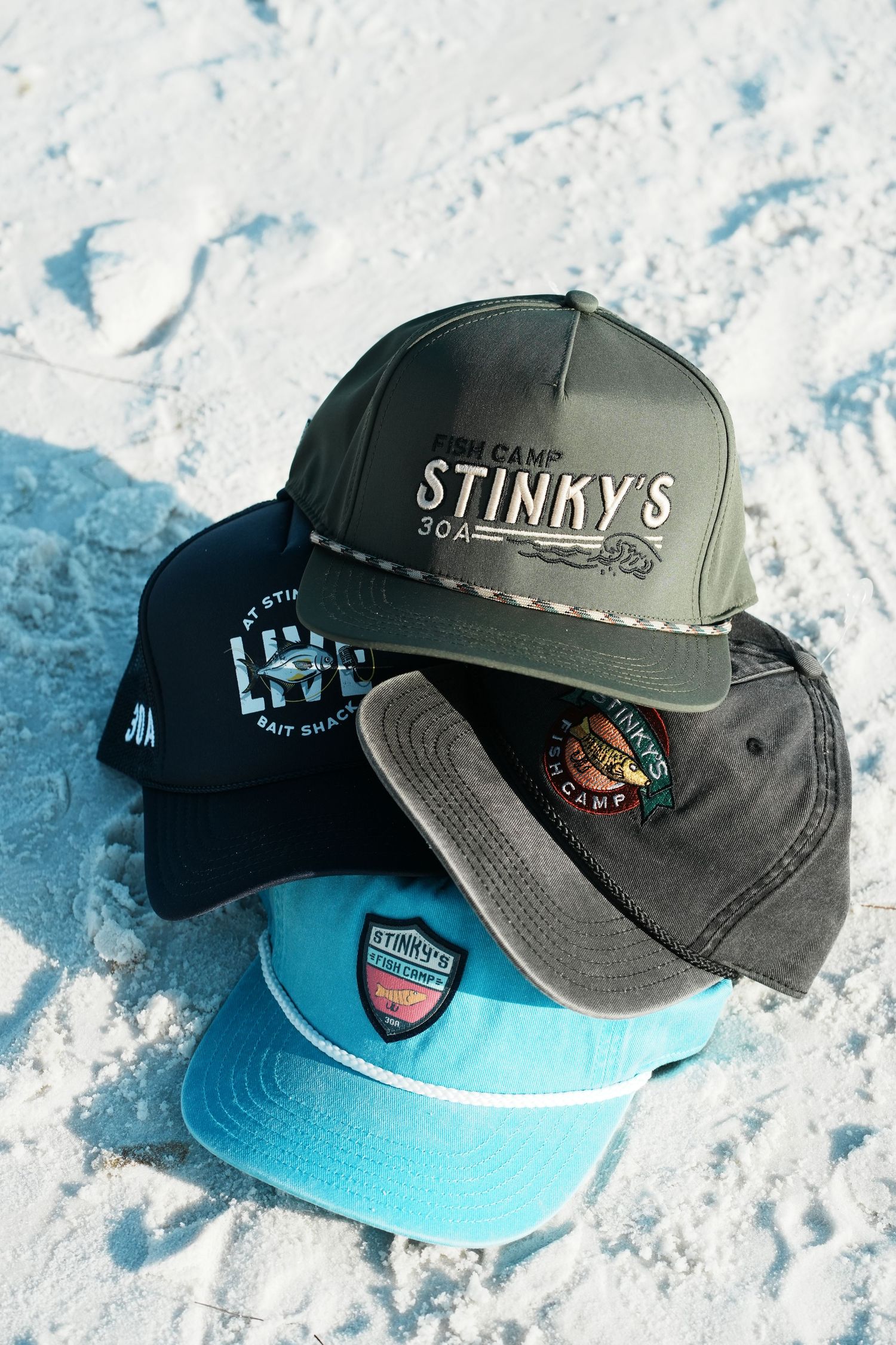 A collection of four branded baseball caps from Stinky's Fish Camp in various colors displayed on a sandy surface.
