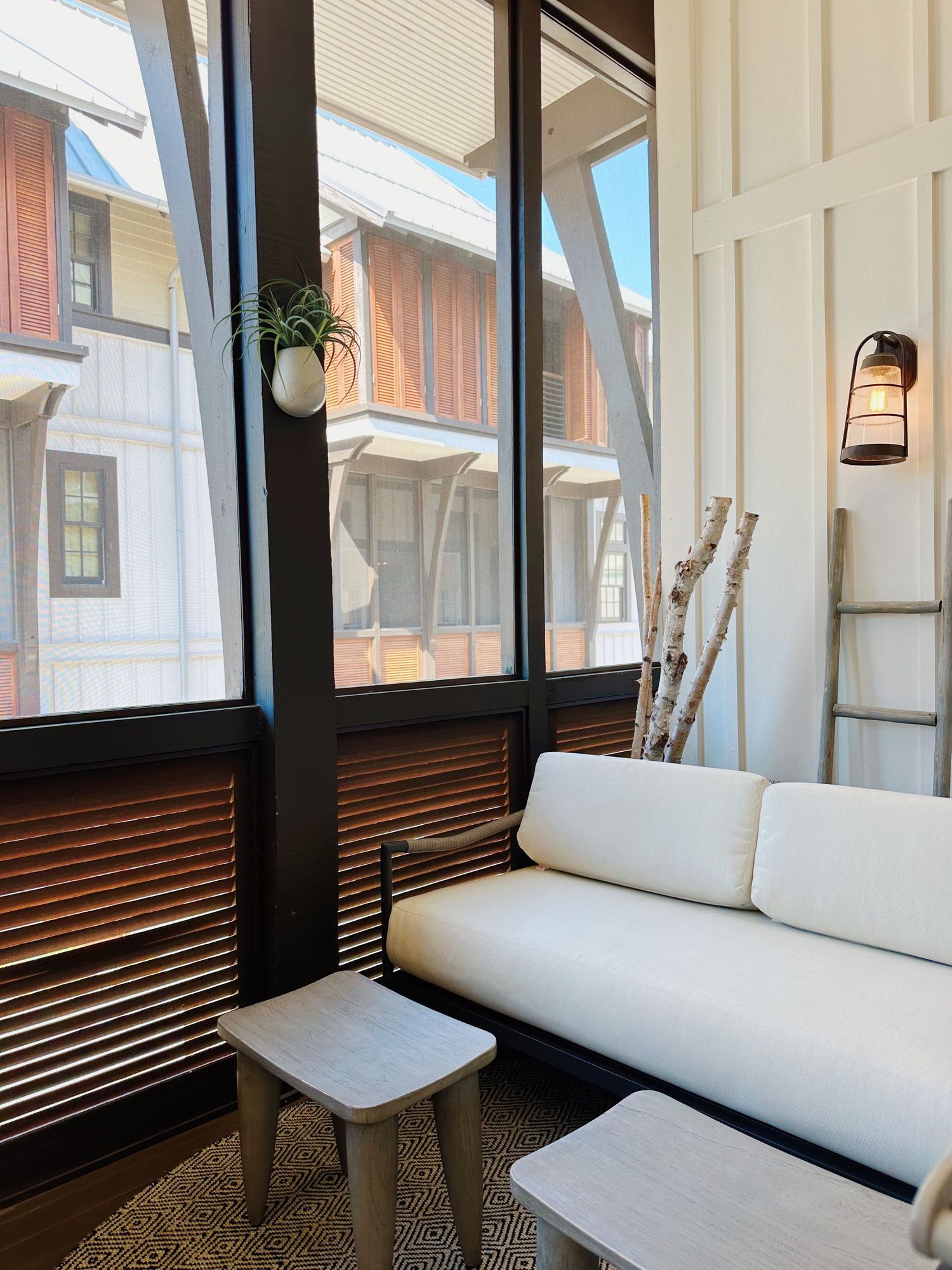 Modern sunroom with white cushioned seating, wooden slatted walls, and large windows overlooking exterior buildings.