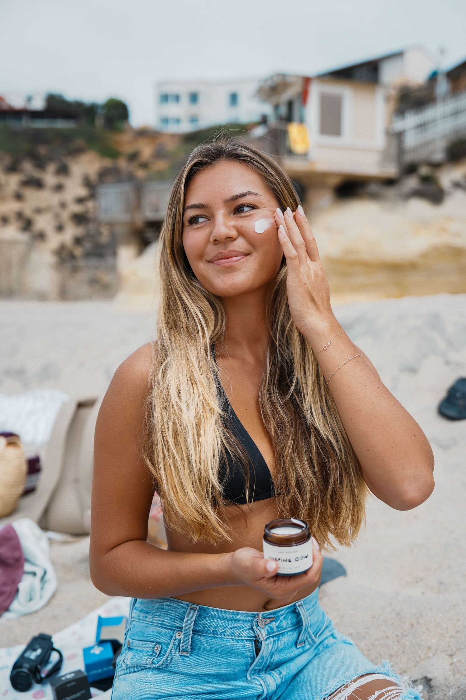 Woman in denim shorts sits on beach sand while enjoying coffee and smiling at sunset.