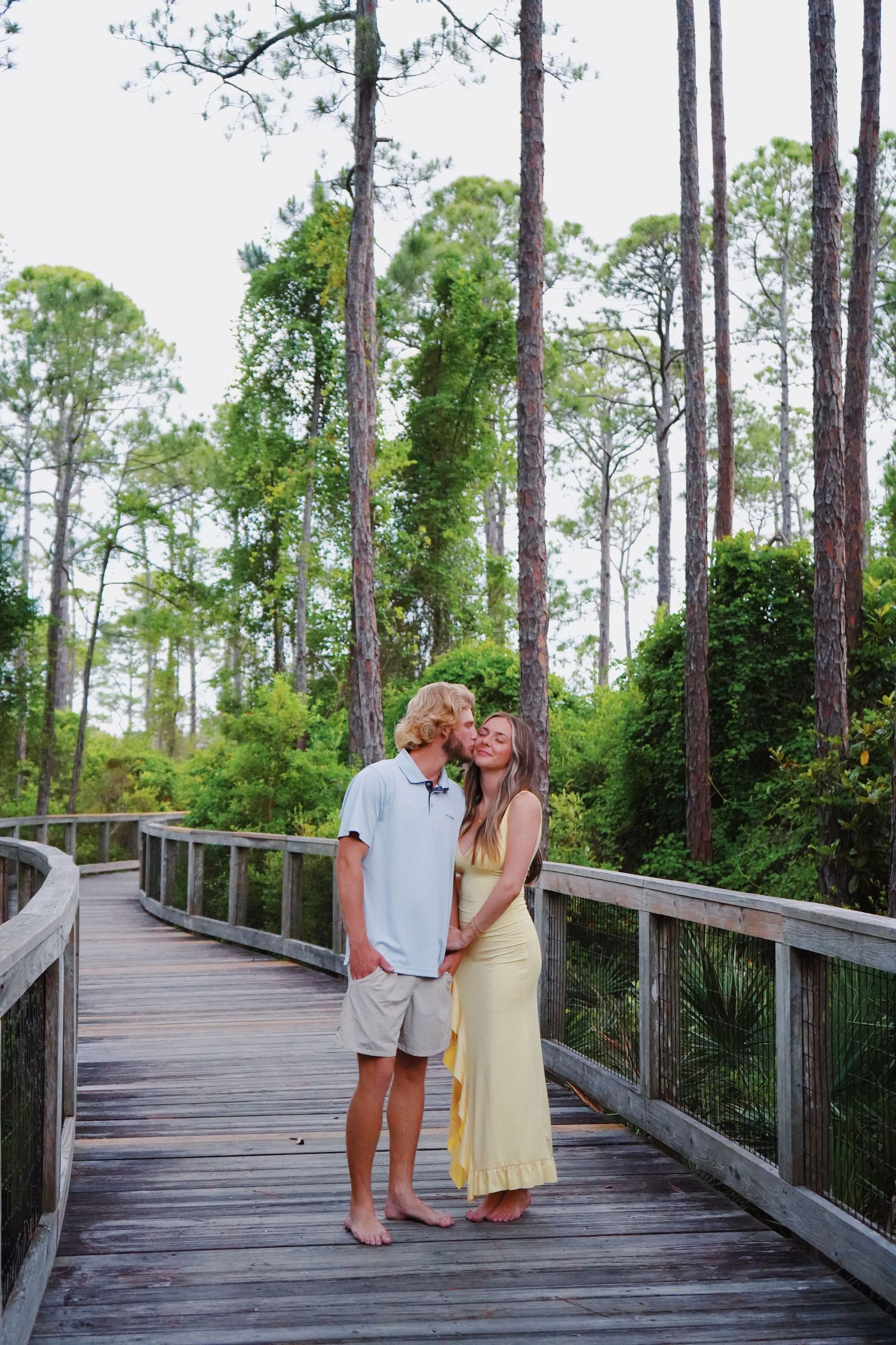 A couple stands together on a wooden boardwalk surrounded by tall pine trees and lush greenery.