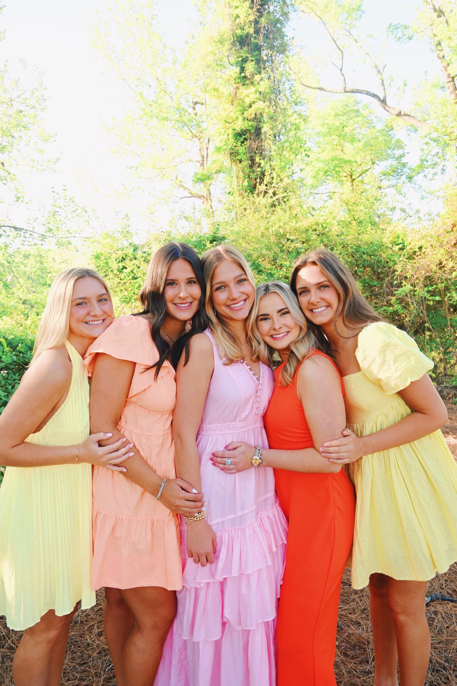 Group of friends in colorful summer dresses pose together outdoors in bright sunlight.