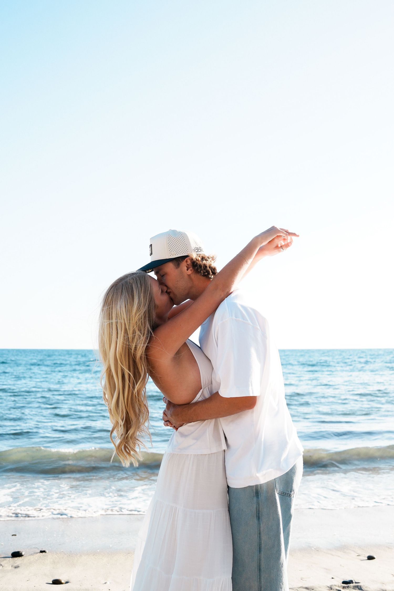 Couple kissing romantically on a sunny beach, woman in white dress with long blonde hair, man in white shirt and cap.