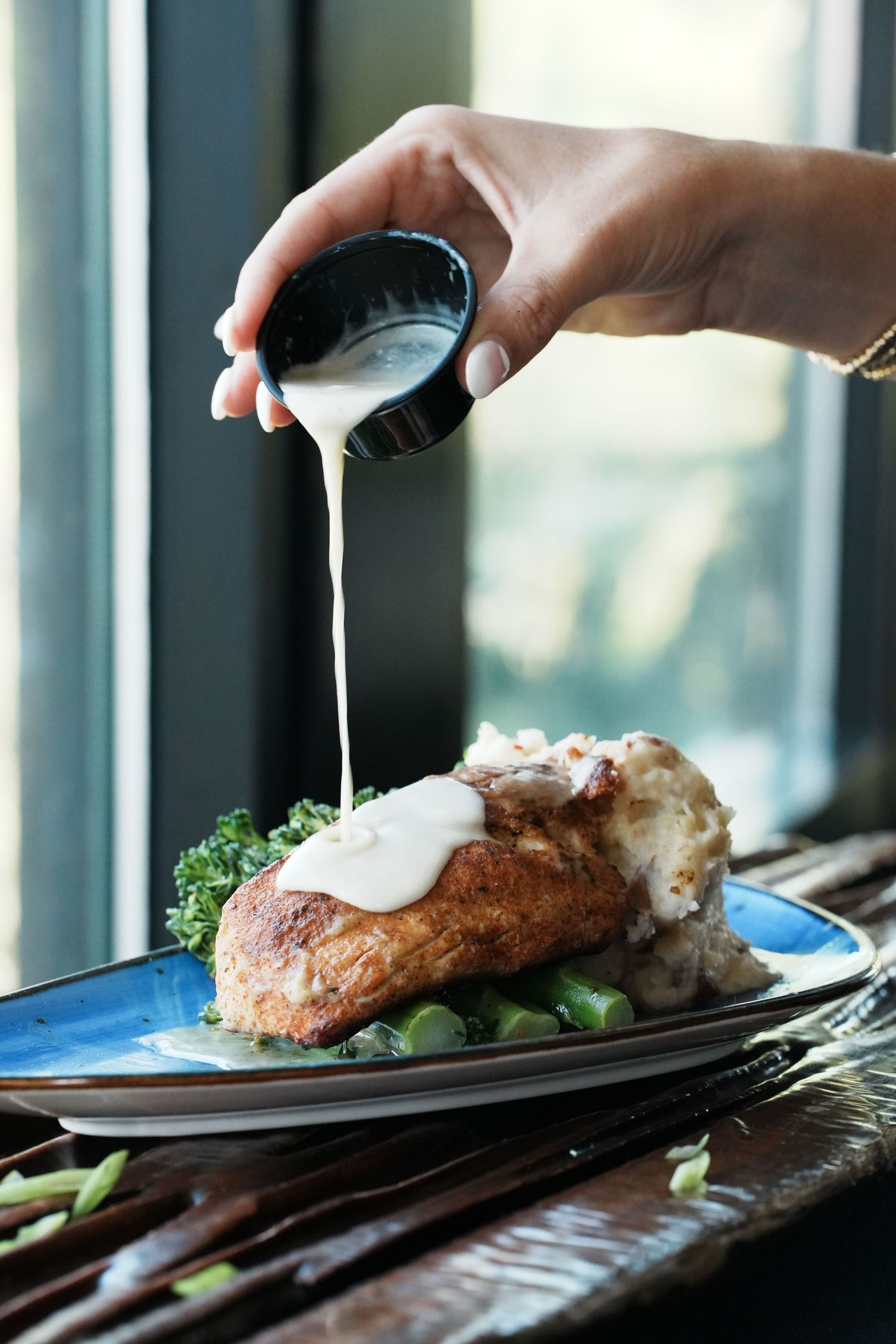 Creamy white sauce being poured over a grilled meat dish with vegetables on a blue plate.