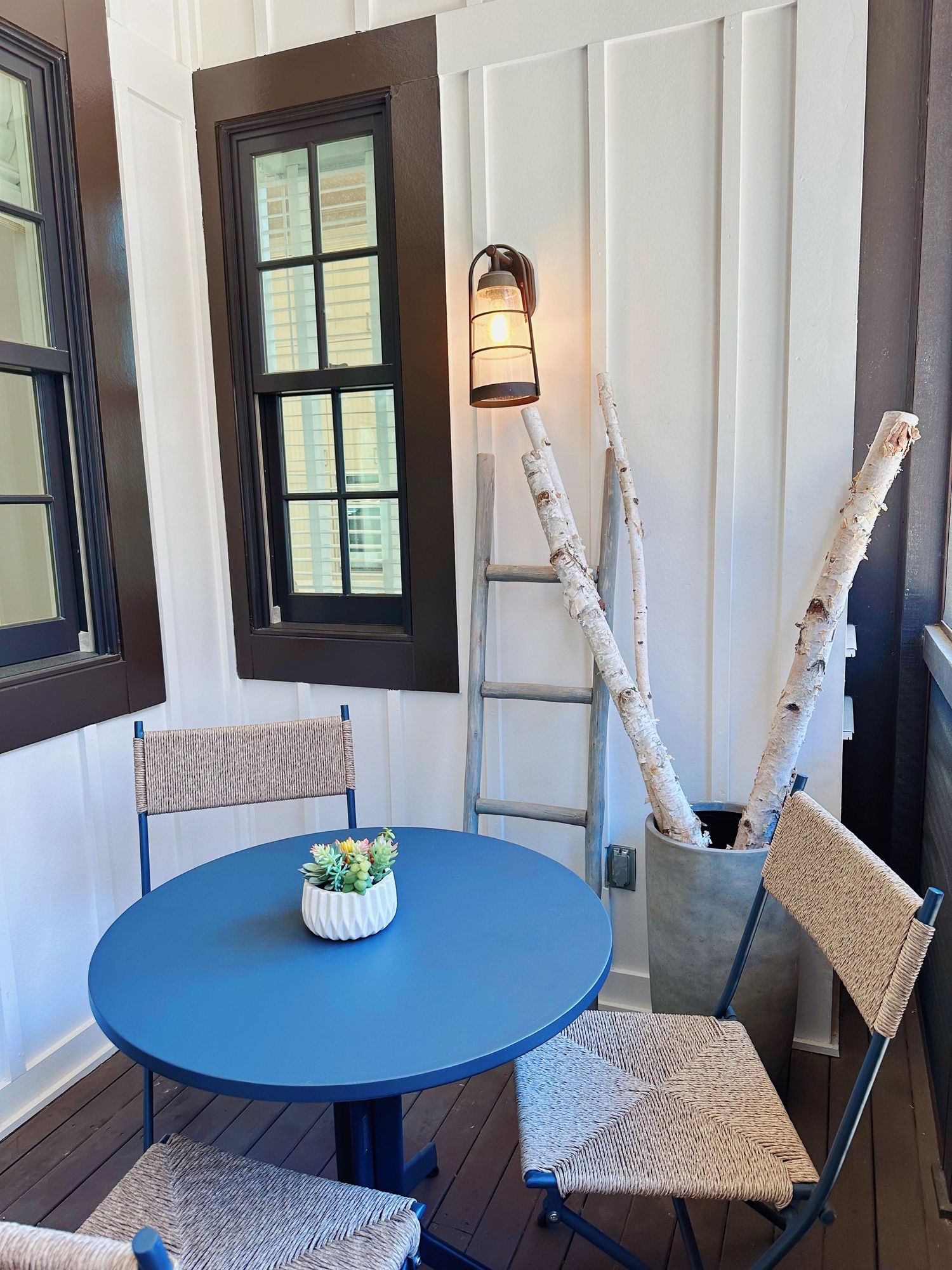A cozy porch corner with a round blue table, cushioned chairs, birch branches, and a lantern-style wall sconce.