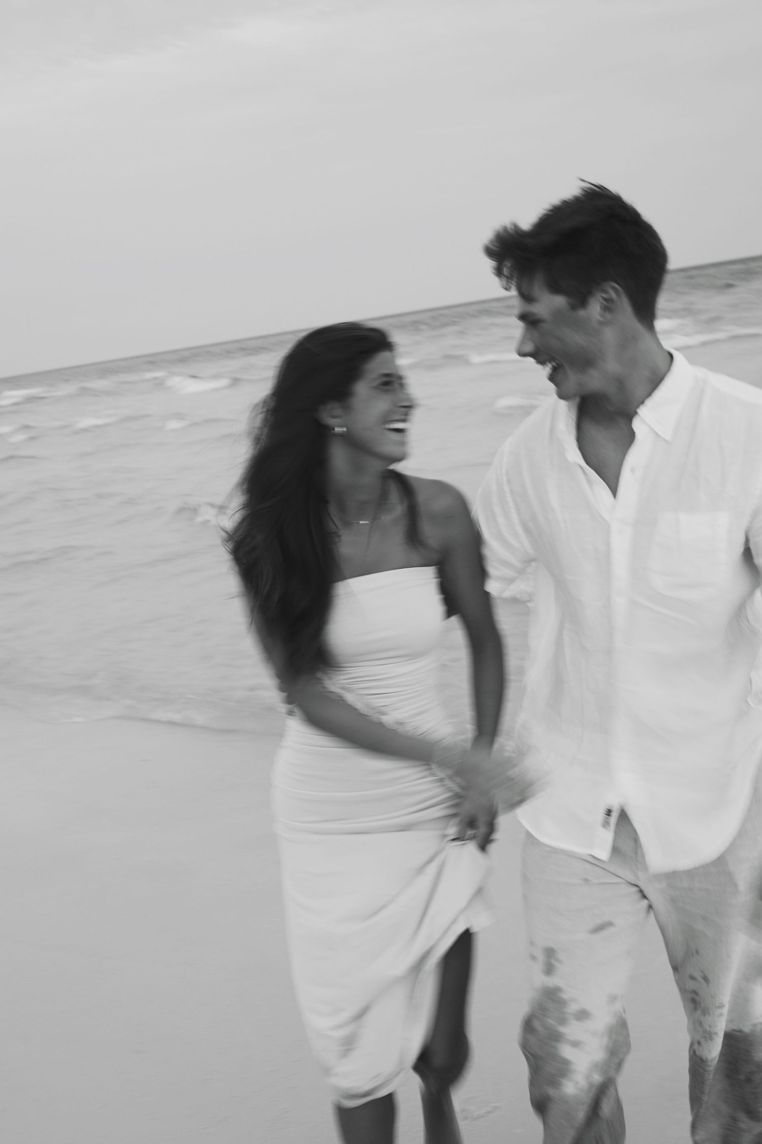 Romantic couple walking and embracing on beach wearing white clothes against ocean backdrop in black and white photograph.