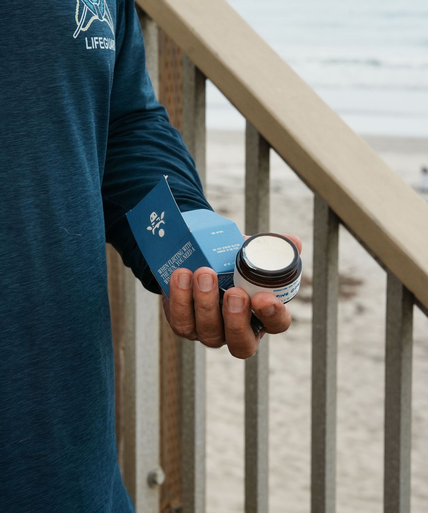 Hands holding a blue-labeled skincare product against a wooden railing with beach scenery.