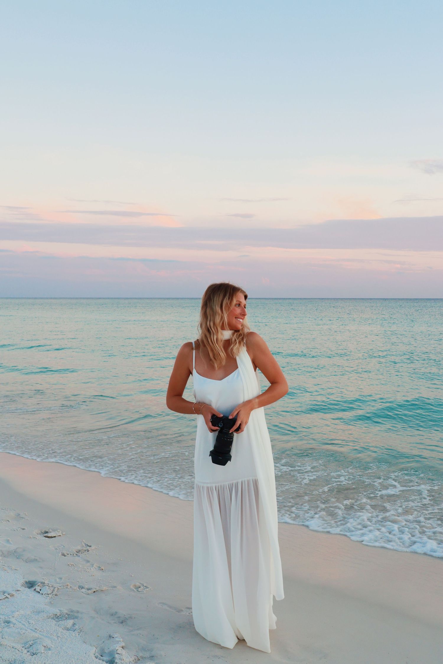 A person in a flowing white dress poses gracefully on a serene beach at sunset with turquoise waters in the background.
