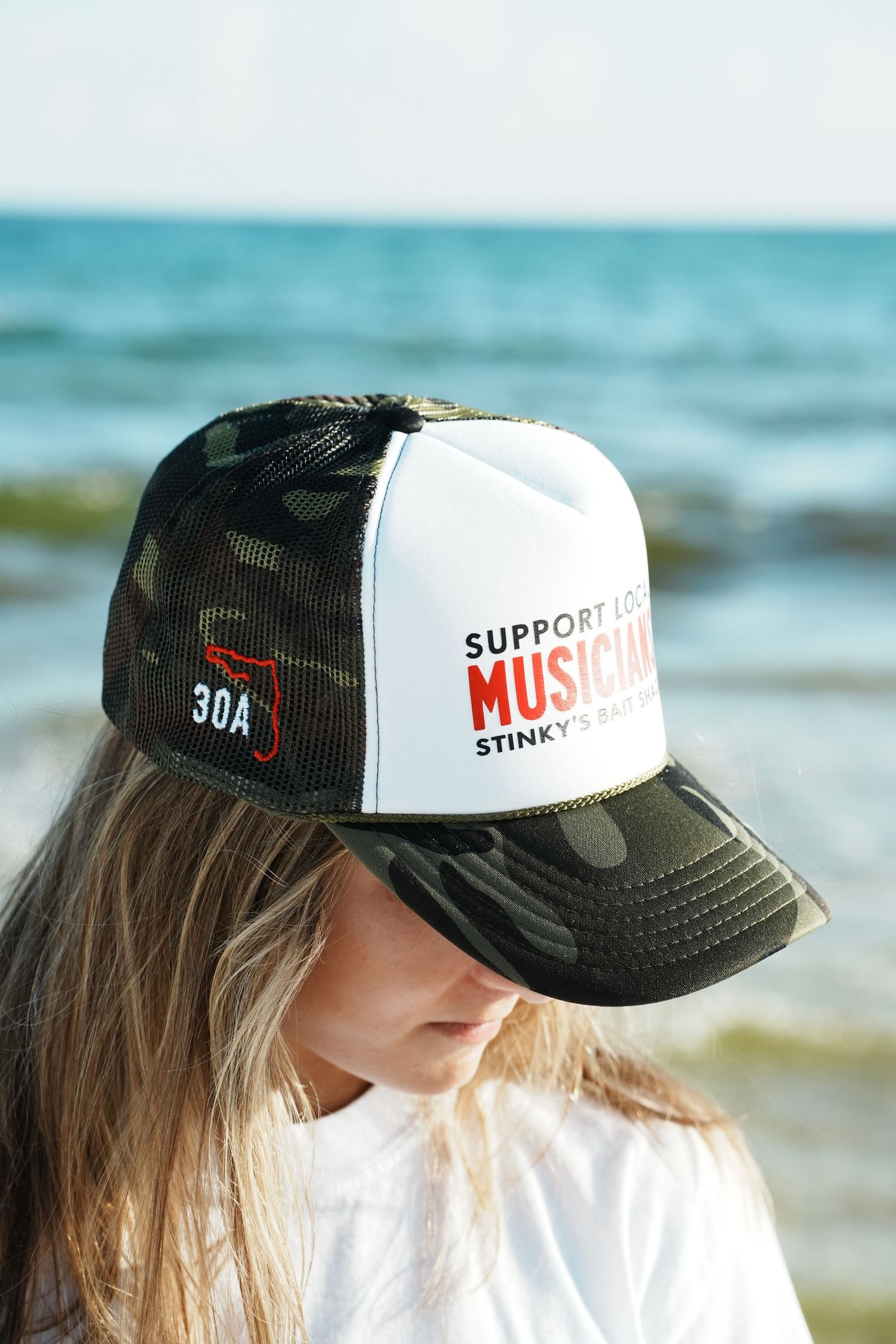 Camouflage and white trucker hat with 'Support Music' text shown against a blurred beach background.