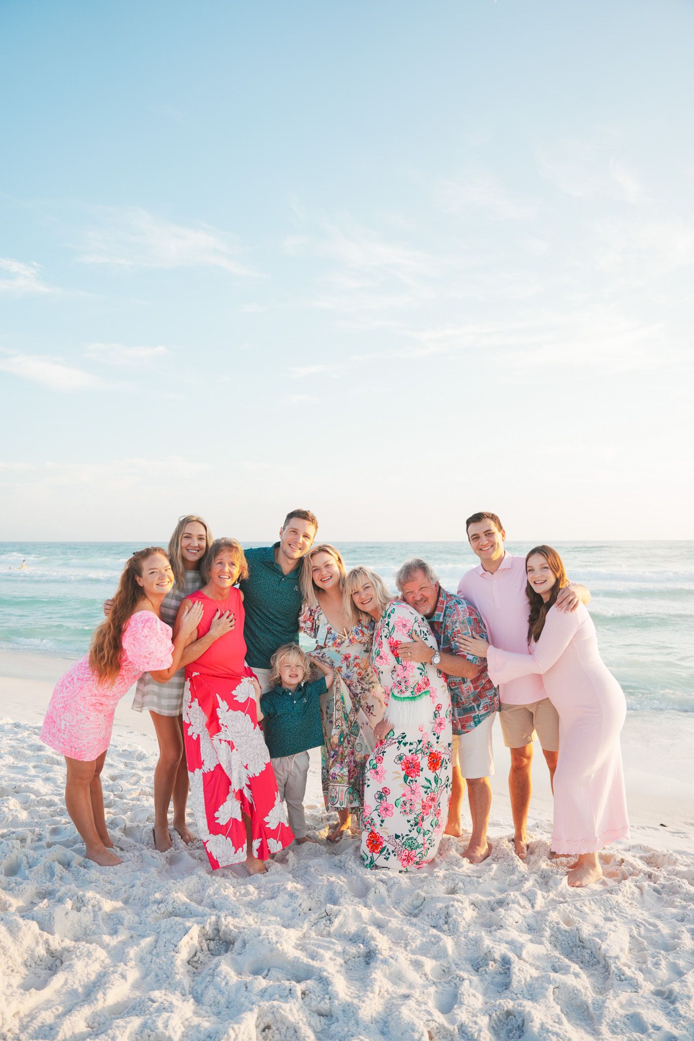 A group in coordinated pink and floral outfits poses together on a sunny white sand beach with ocean waves in the background.