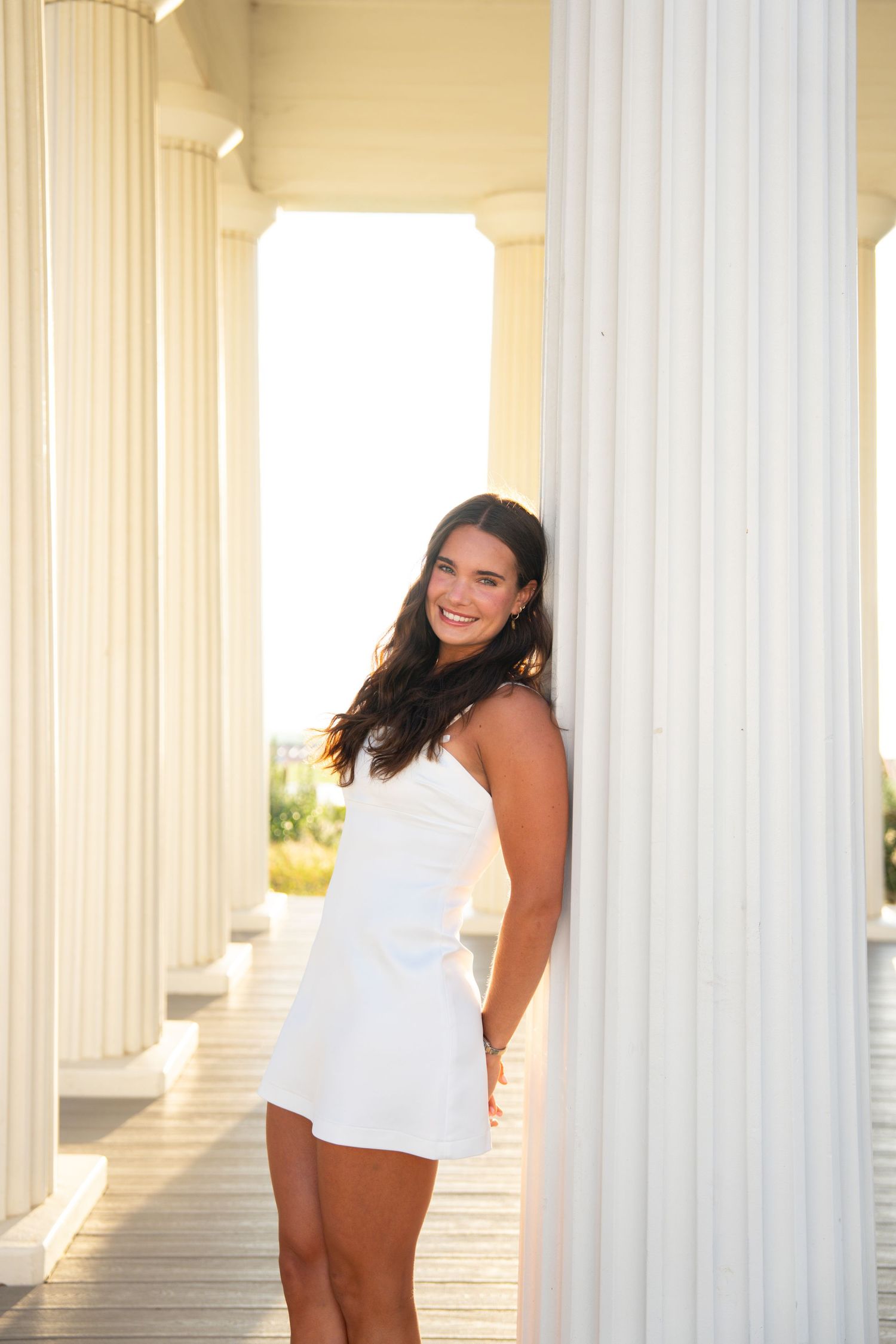 Woman in white dress poses between elegant white columns during a sunny outdoor photoshoot.