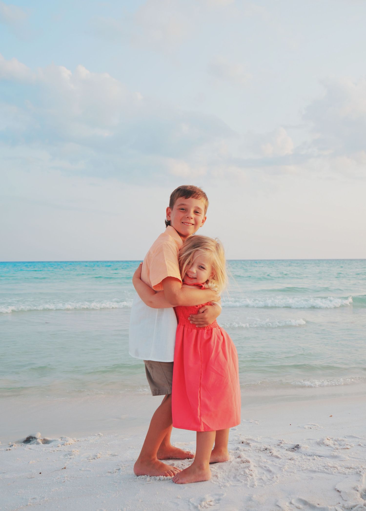 Two children embrace while standing on a white sandy beach with turquoise ocean waves and blue sky in the background.