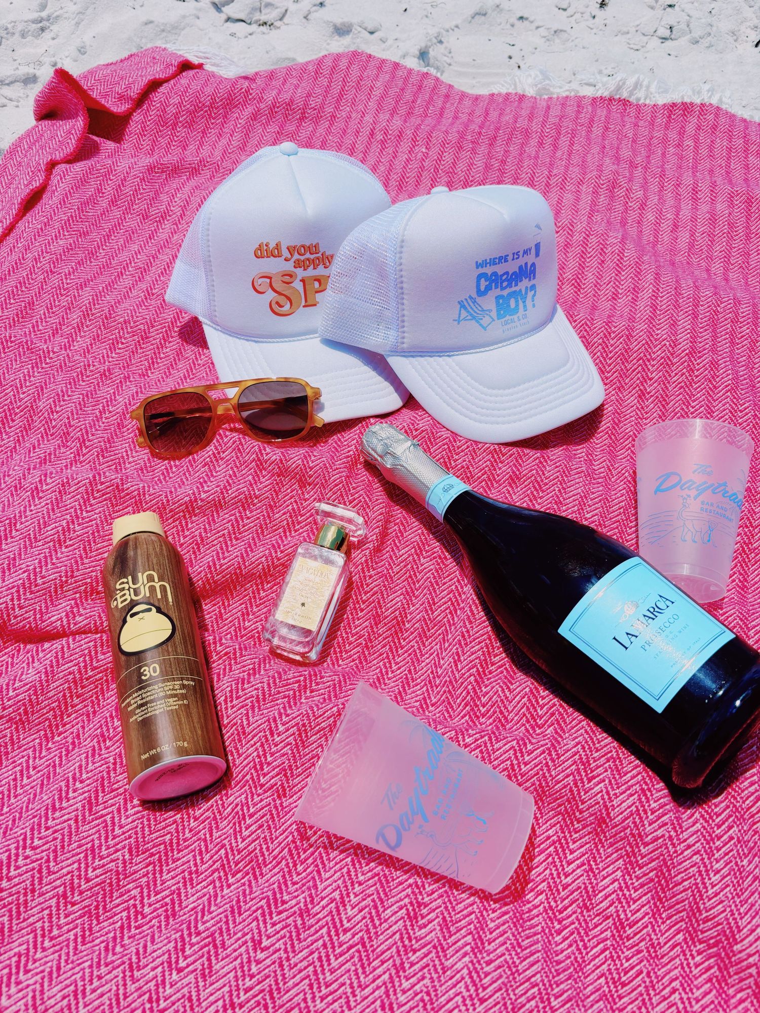 Summer beach accessories including white caps, sunglasses, drinks and bottles laid out on pink towel.