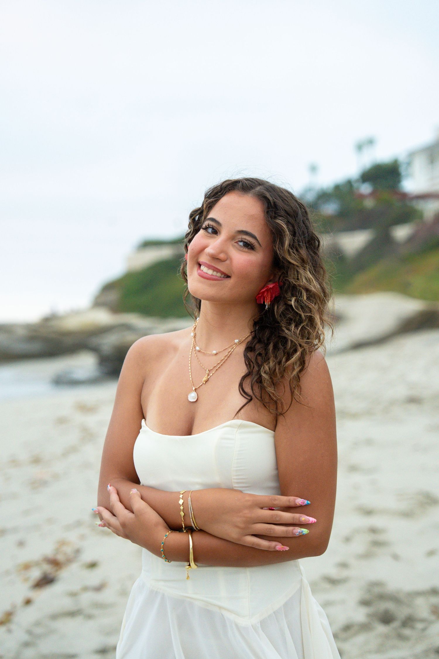 Smiling young woman in white strapless dress with red flower earring posing on sandy beach with rocky cliffs.