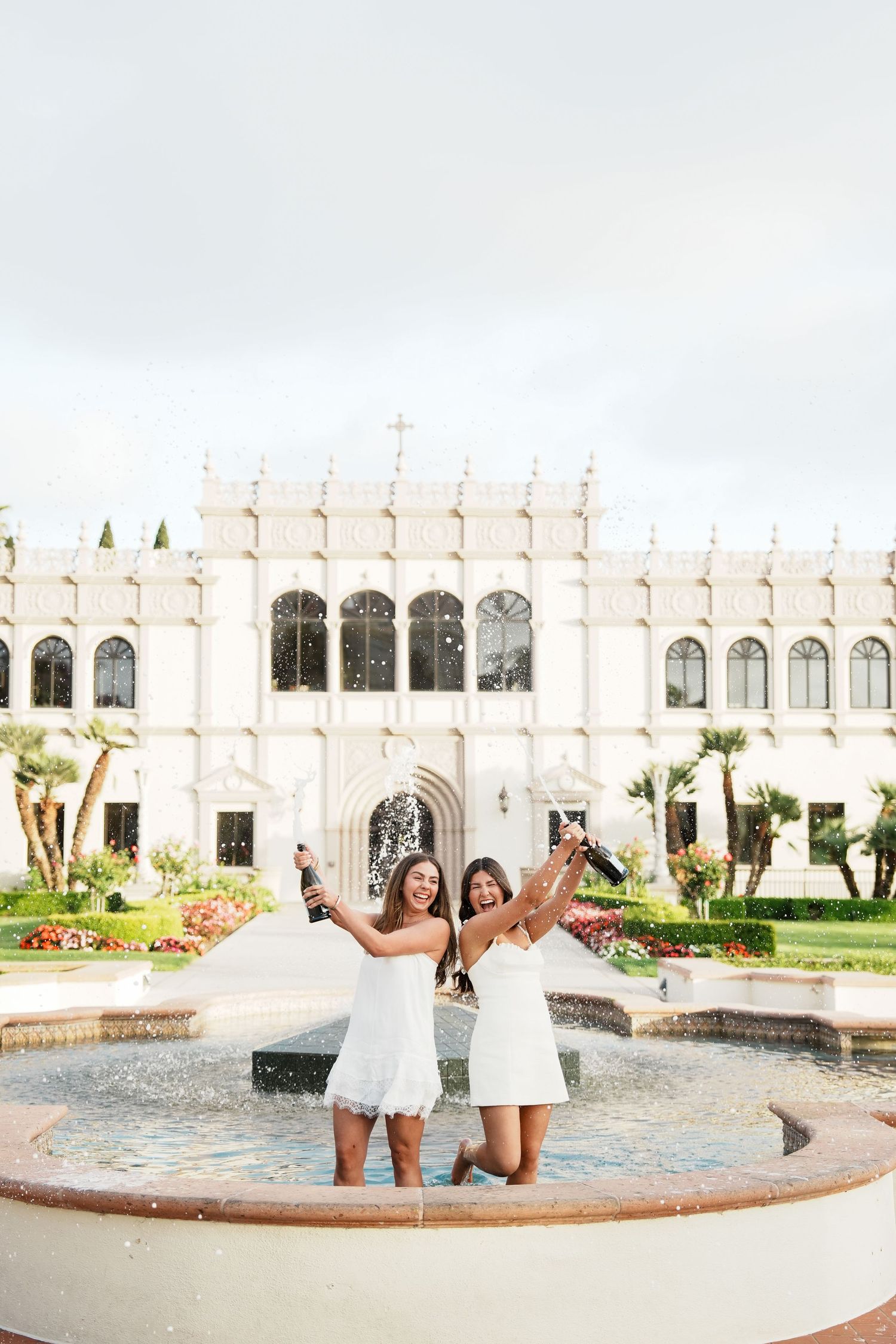 Two women in white dresses celebrate with champagne bottles in a fountain outside an elegant white building.