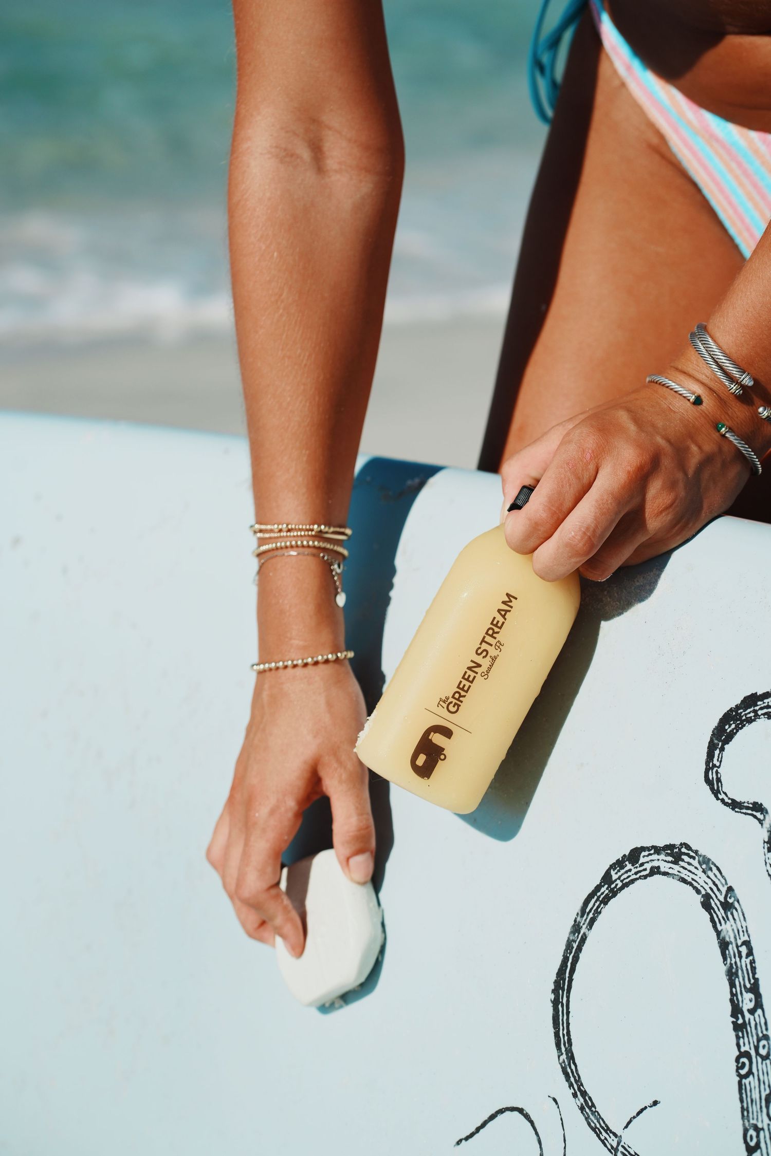 Hand with delicate gold anklets holds a beige surf wax bar over a surfboard at the beach.