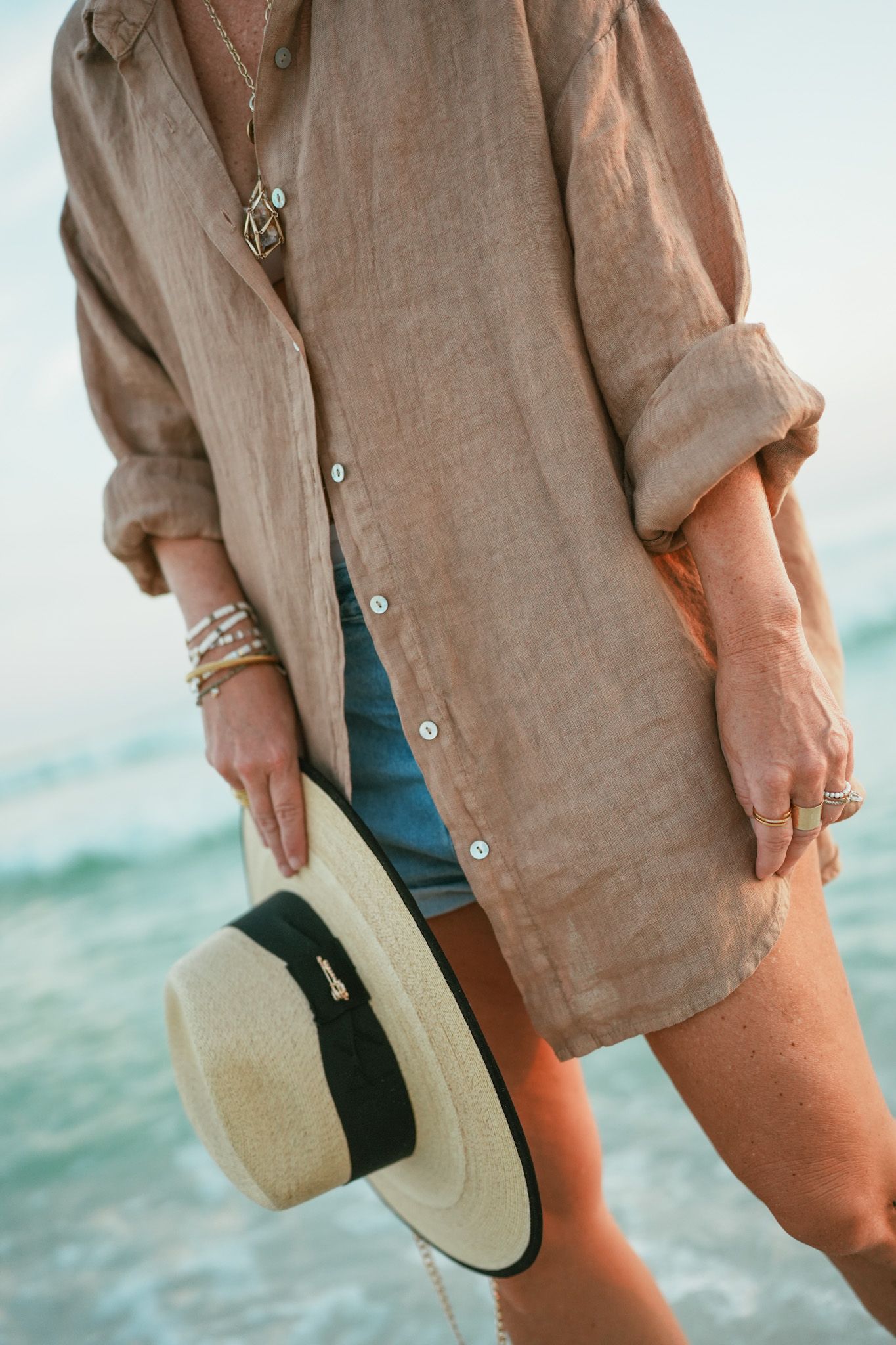 Close-up fashion details of a beige linen shirt and sunhat against an ocean backdrop.