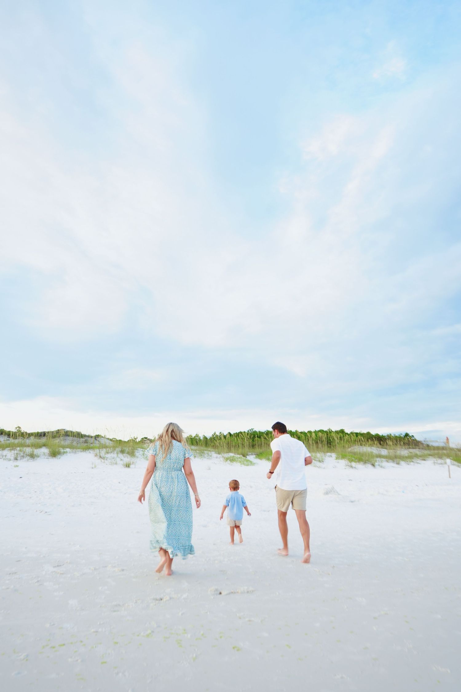 A family walks hand in hand along a beautiful white sand beach with dunes in the background.