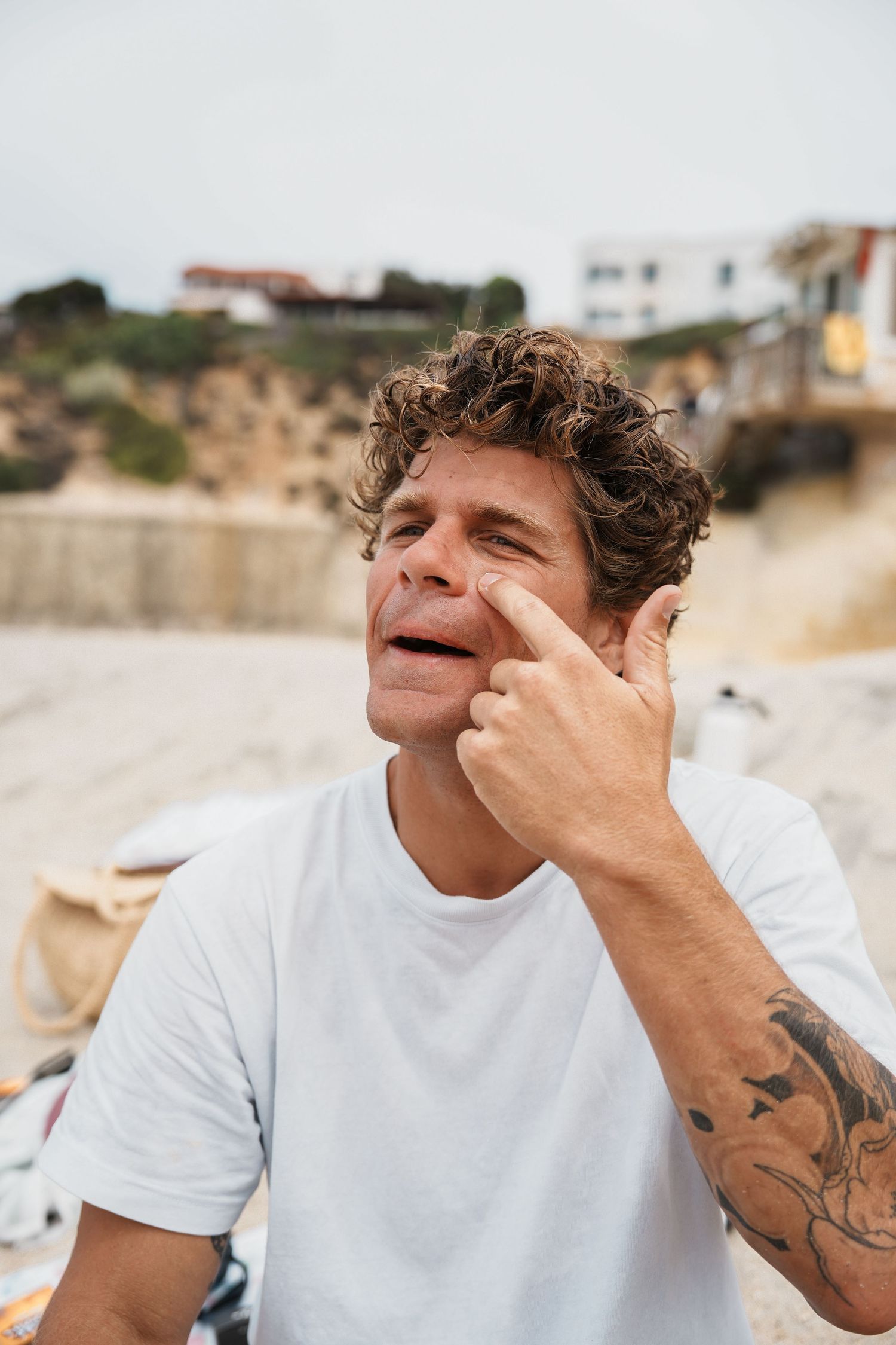 Person wearing white t-shirt with tattoos smiling on a beach with cliffs in background.