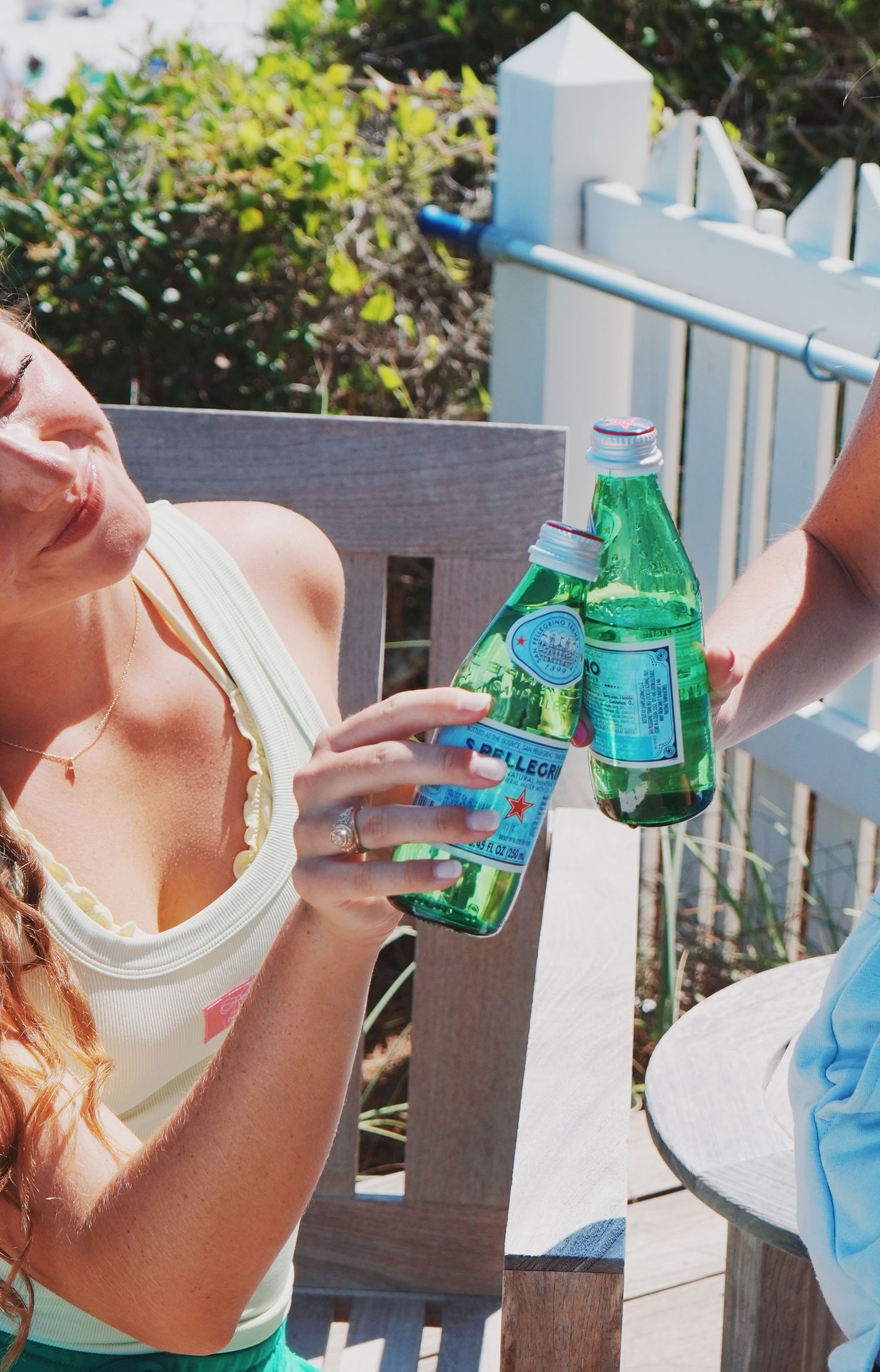 People toasting with glass bottles of Sprite soda on a sunny outdoor patio with white picket fence in background.