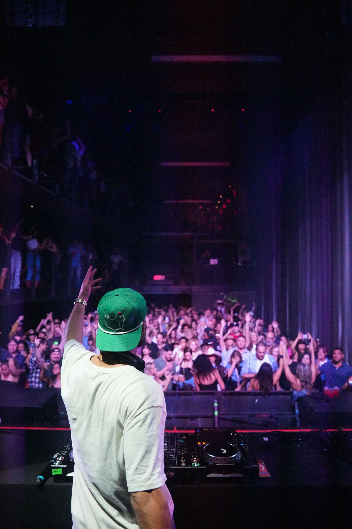 DJ performs at a nightclub with an energetic crowd in the background illuminated by purple lighting.