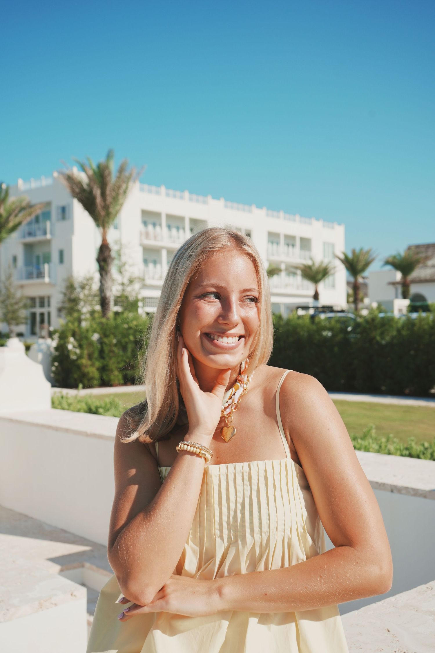 Person in a cream-colored fringe dress smiles at a sunny resort with white buildings and palm trees in the background.
