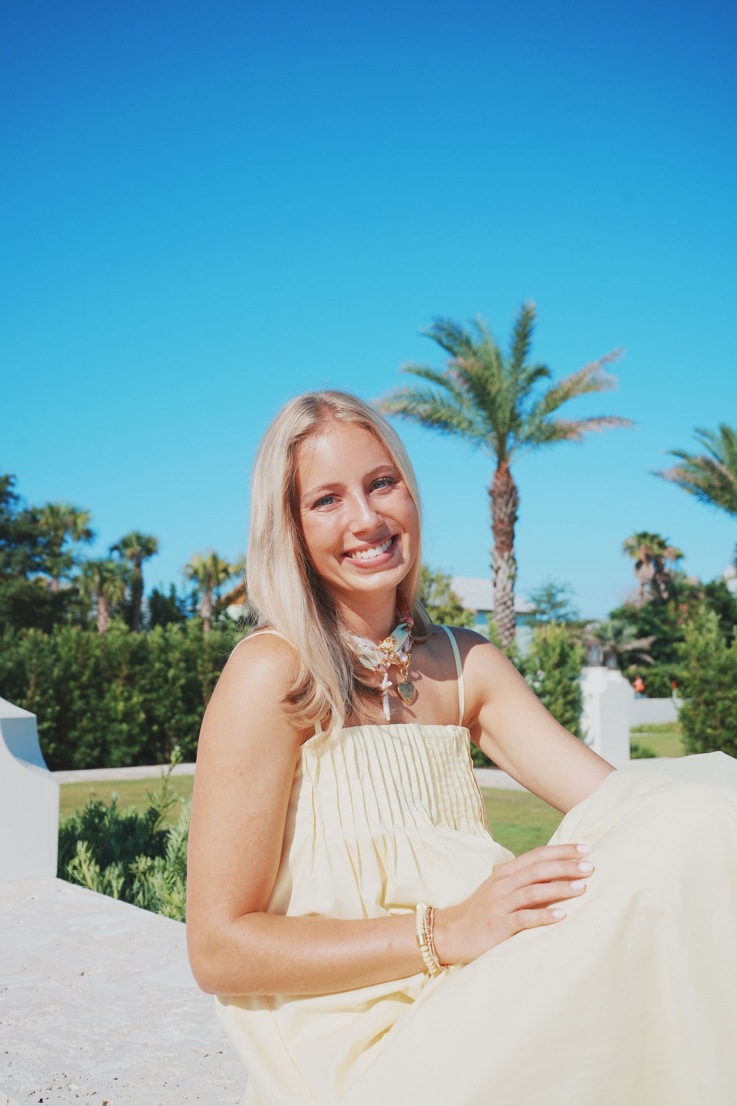Person relaxing on a white lounge chair under palm trees and bright blue sky on a sunny summer day.