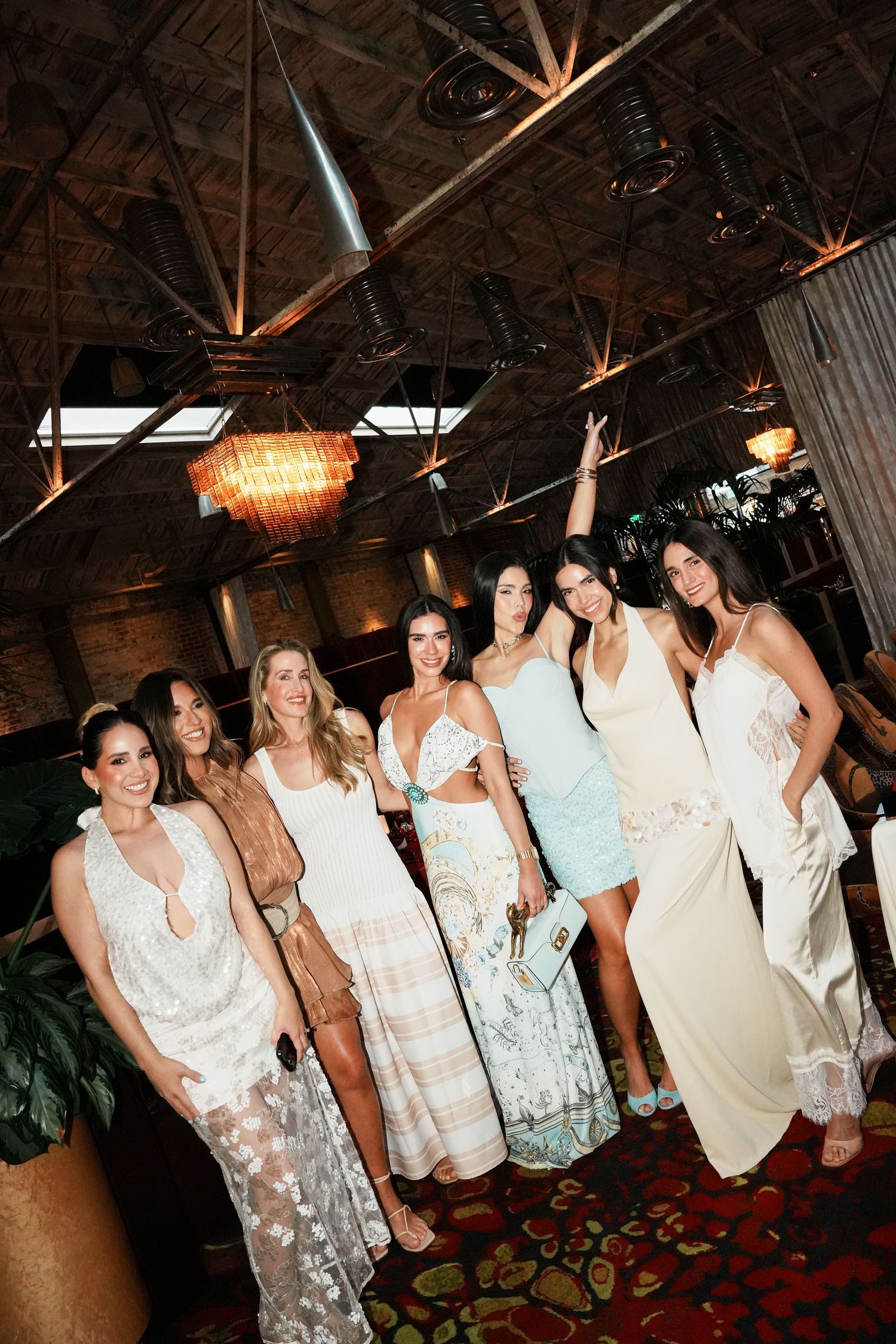 Group of seven women in elegant white and pastel dresses posing together at an upscale indoor venue.