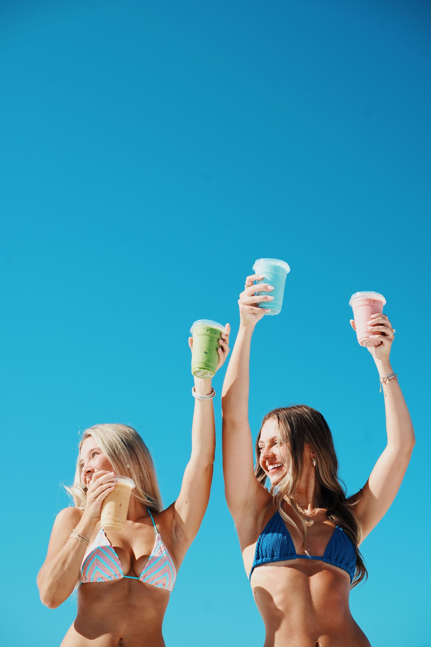 Two women in bikinis raise cold drinks in celebration against bright blue summer sky.