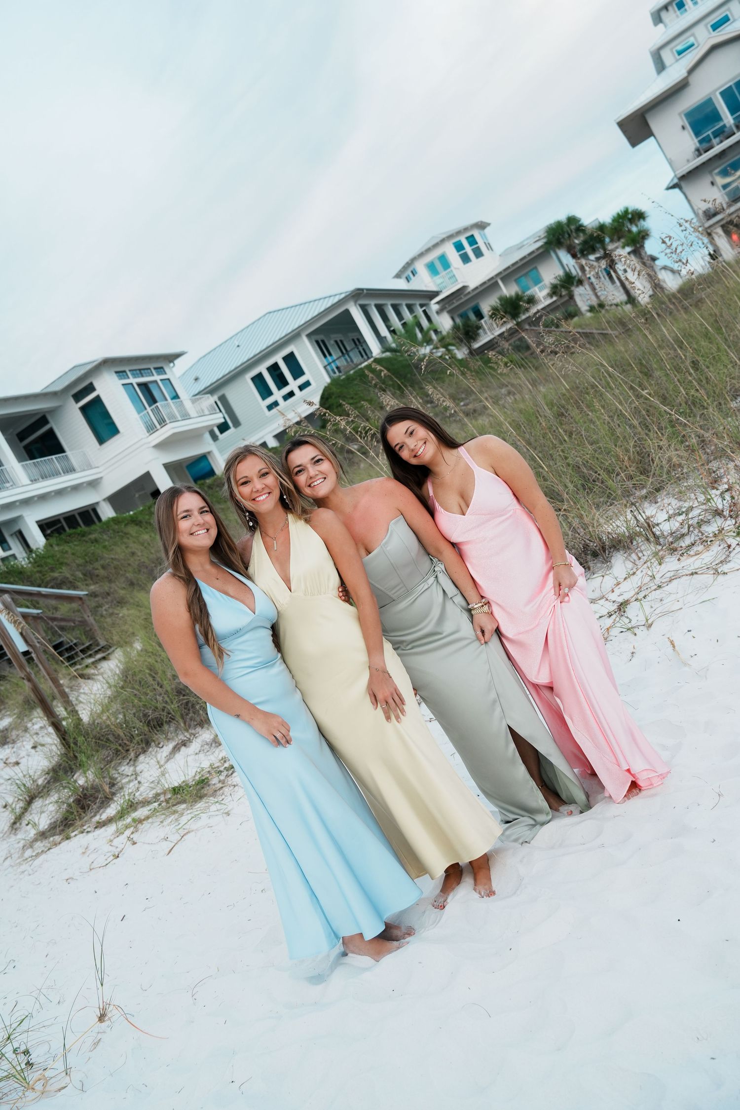 Group in pastel dresses posing together on white sand beach with coastal homes in background.