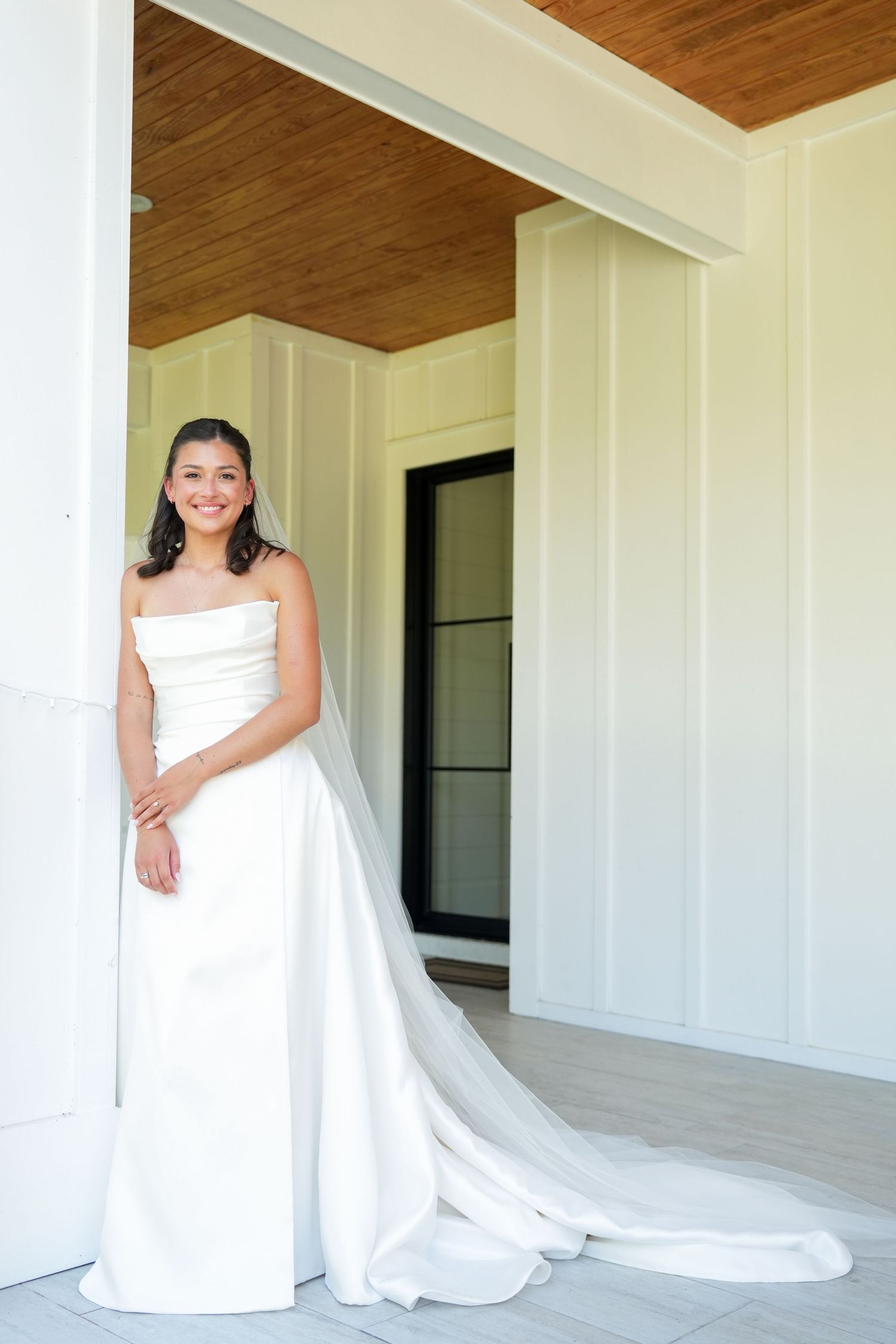 Smiling bride in a strapless white gown with a long train poses by a white column on an elegant porch.