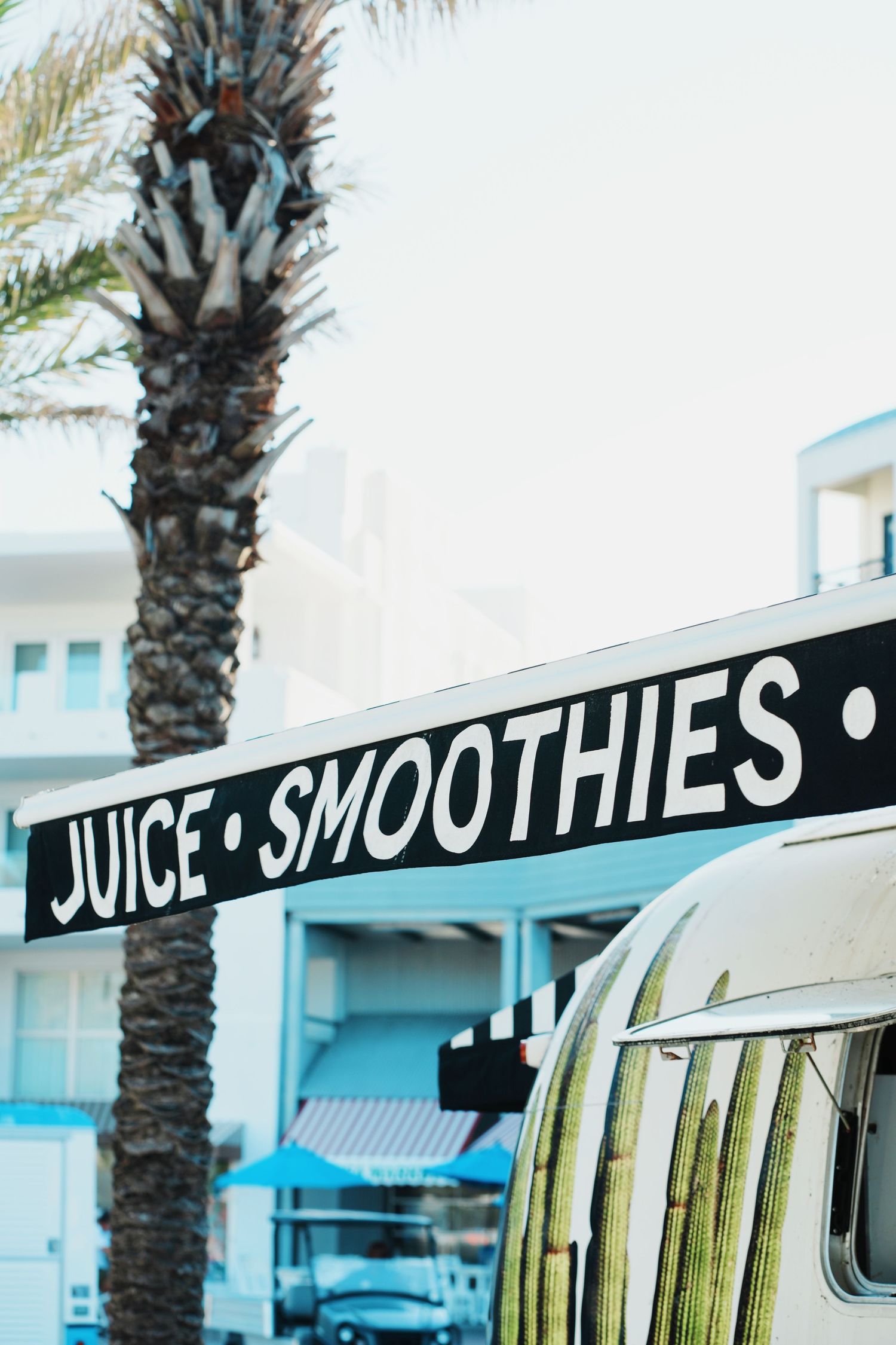 A vintage-style juice and smoothies sign hangs near a palm tree against a beachy blue building backdrop.
