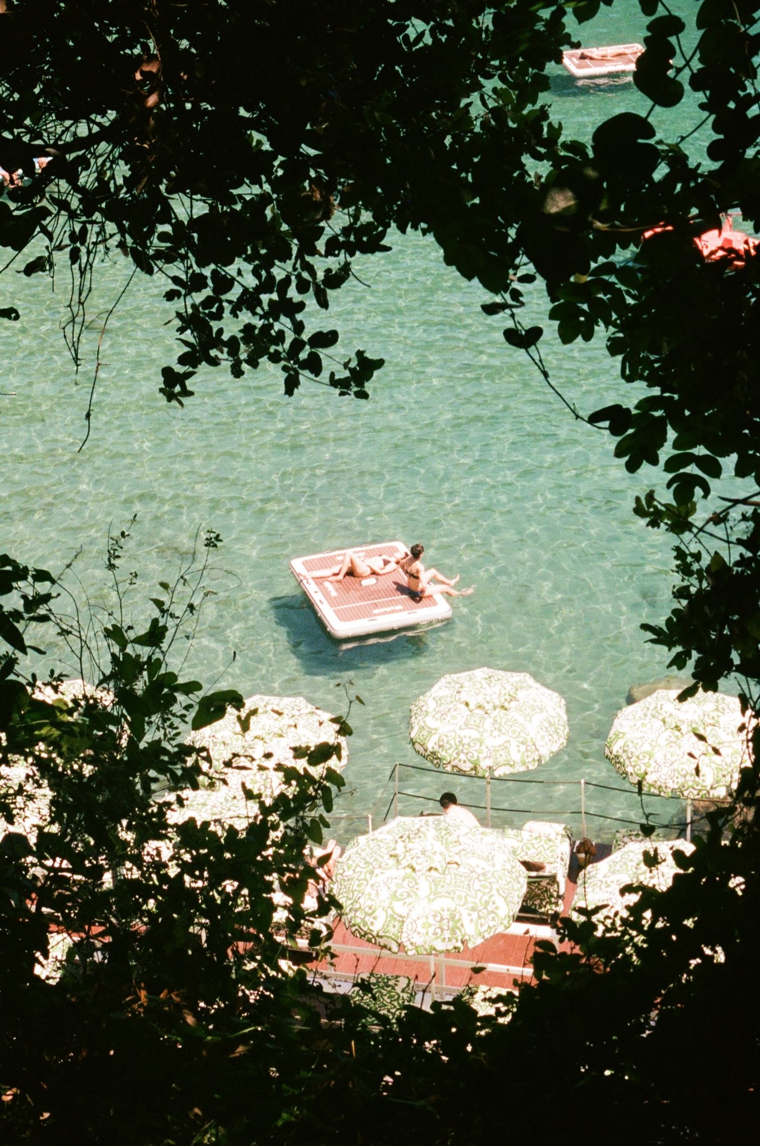 Aerial view of person sunbathing on floating raft in turquoise water, with white beach umbrellas on dock below.