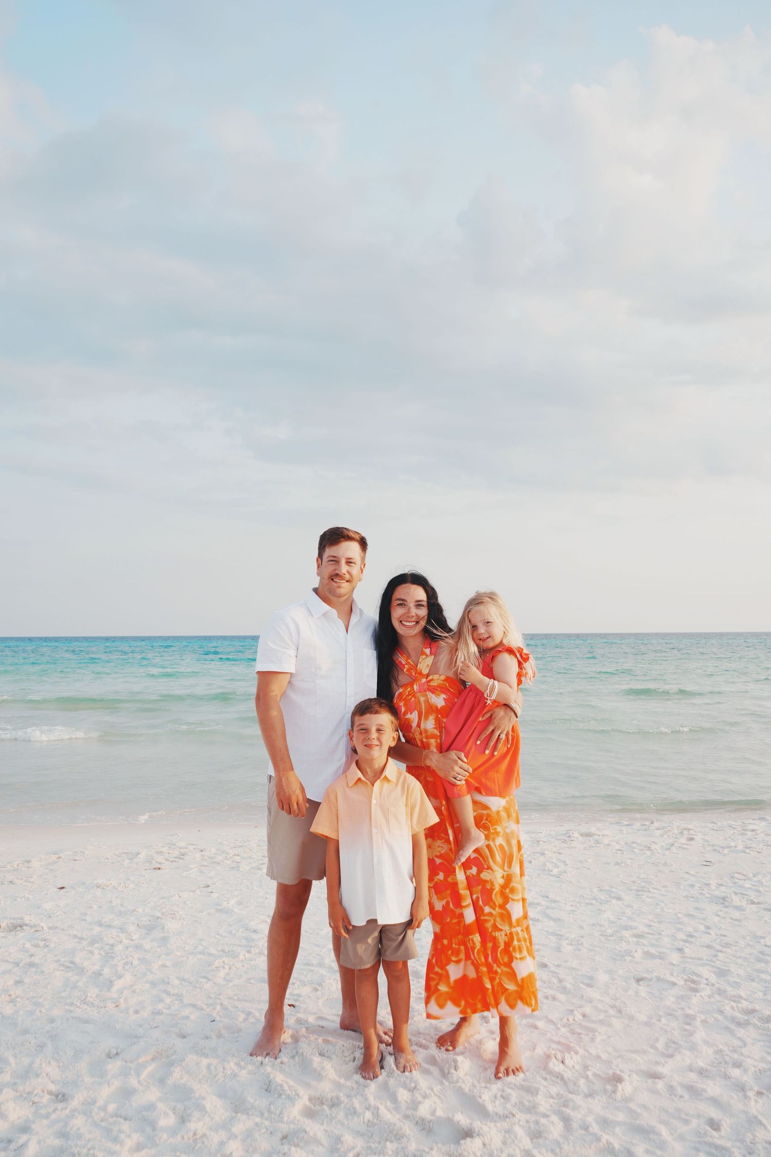 A family of four stands together on a white sandy beach with turquoise waters and soft clouds in the background.