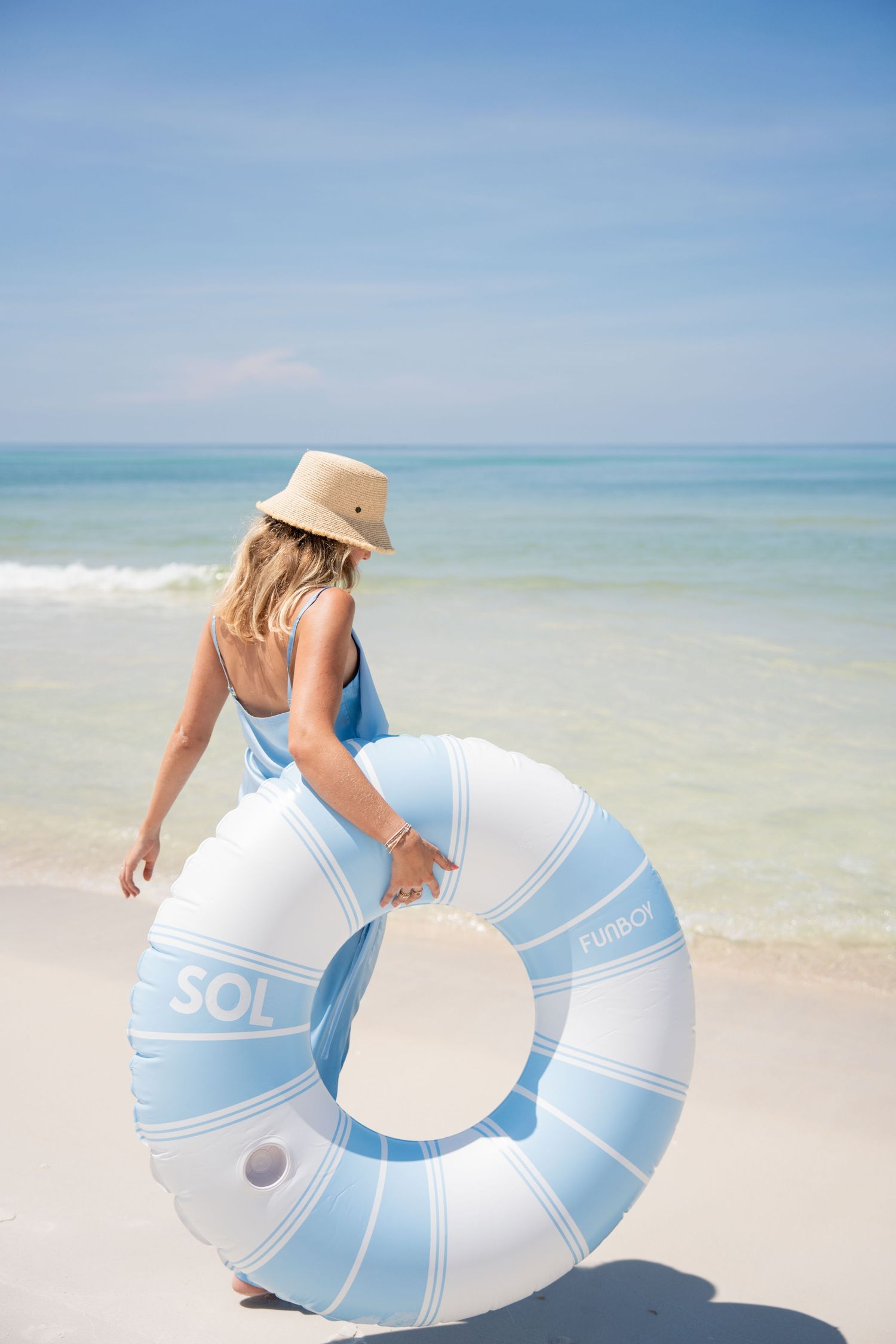 Woman in blue dress and sun hat carrying a light blue SOL inflatable pool ring on a sunny white sand beach.