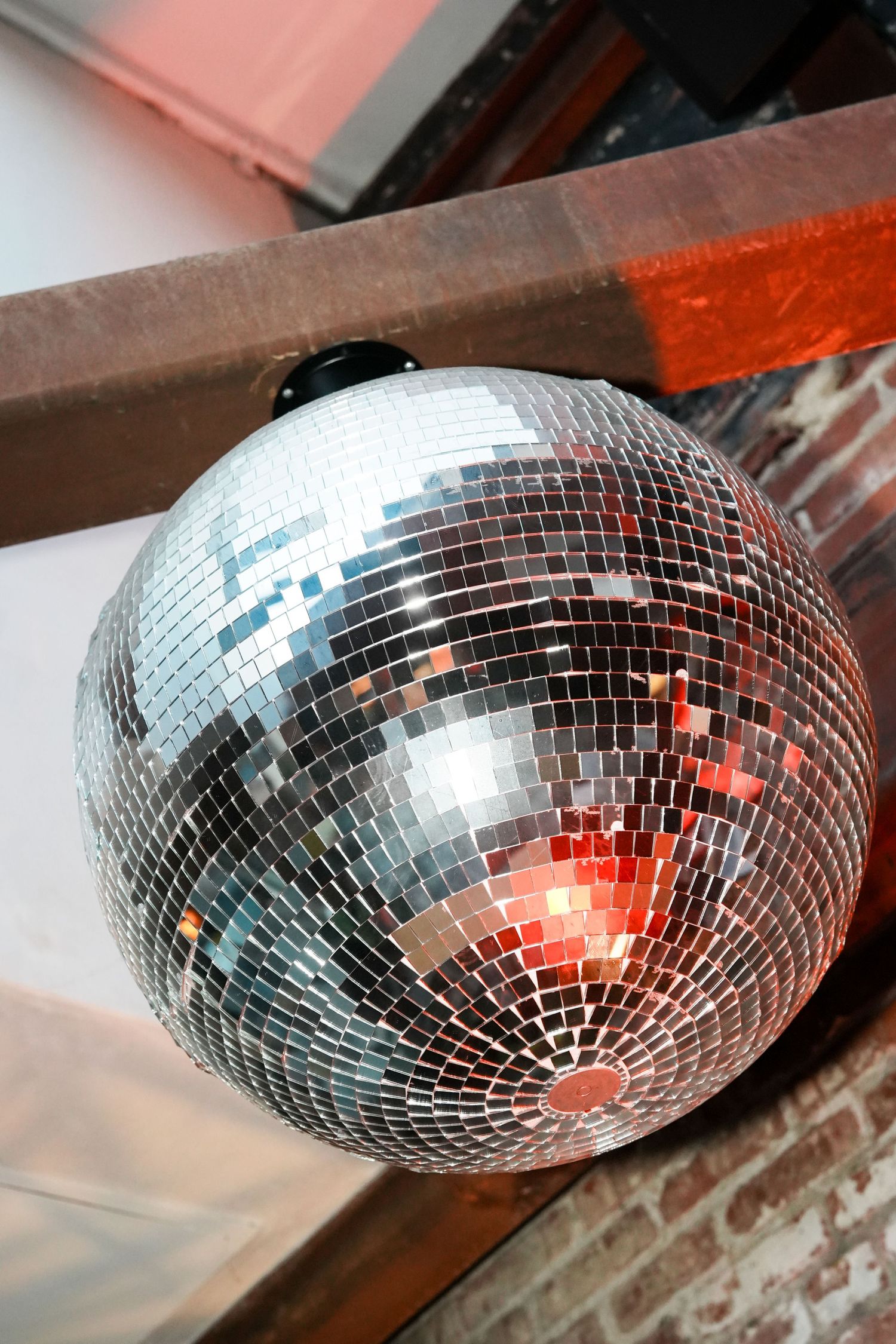 A large silver mirror disco ball hanging from wooden beams reflecting colorful lights at an indoor venue.