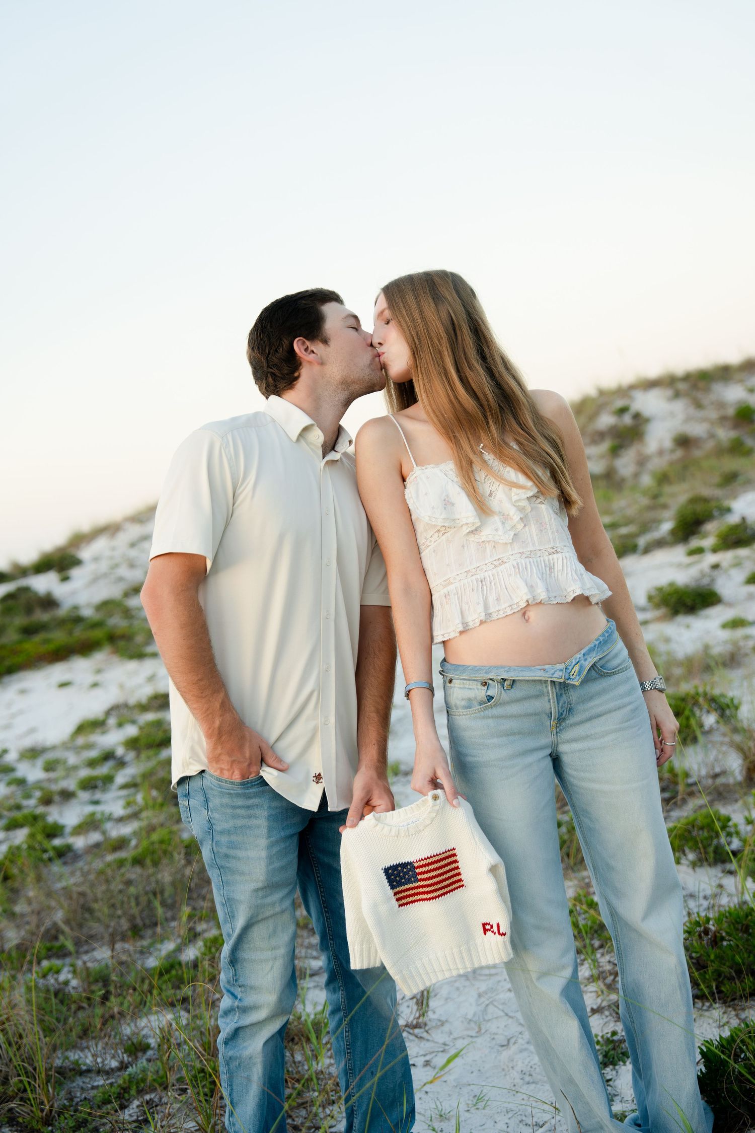 Couple kissing on sandy beach dunes, woman holding American flag tote bag, both wearing jeans and white tops.