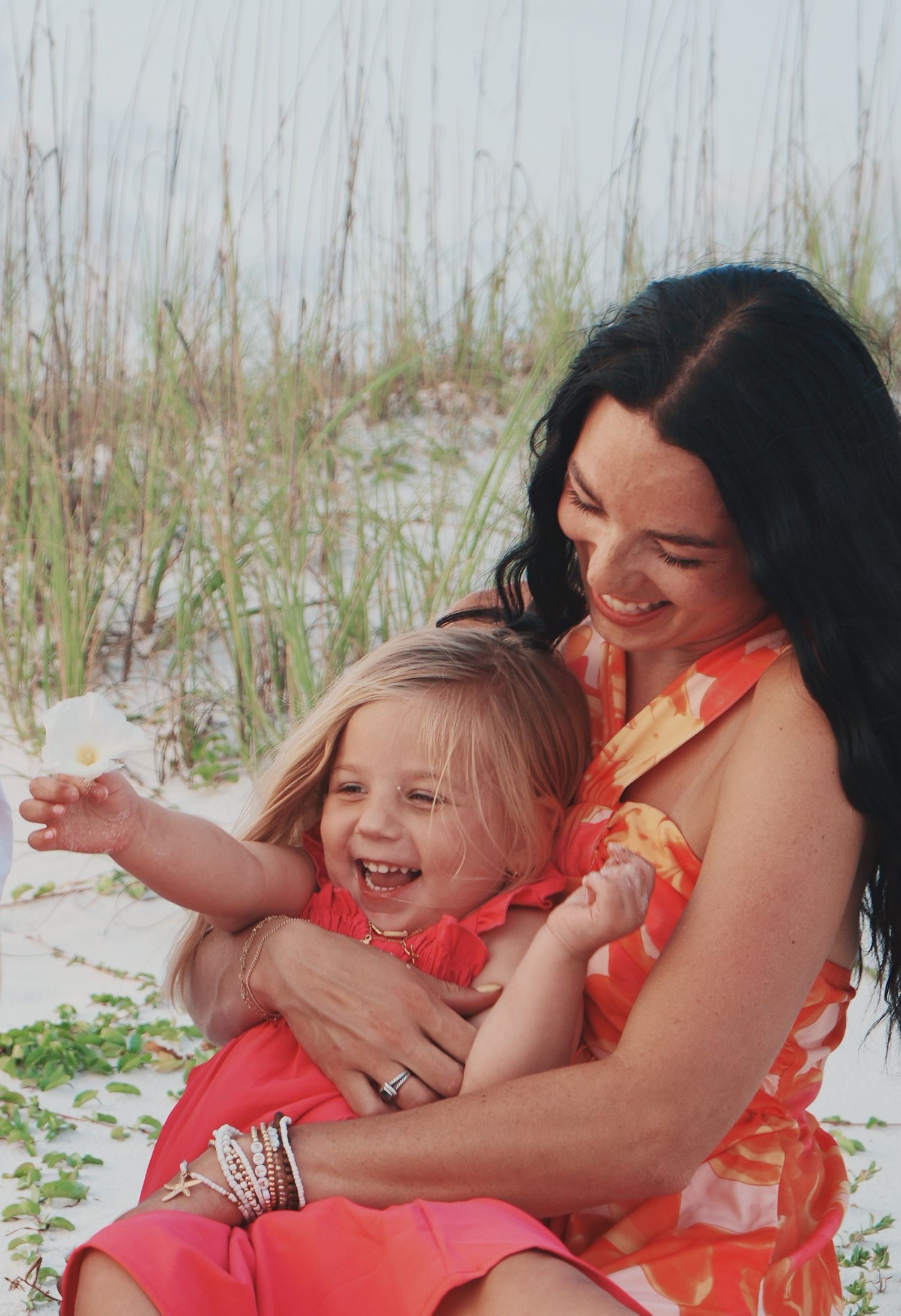A joyful moment shared between parent and child in coral dresses while sitting on white sand with beach grass in background.