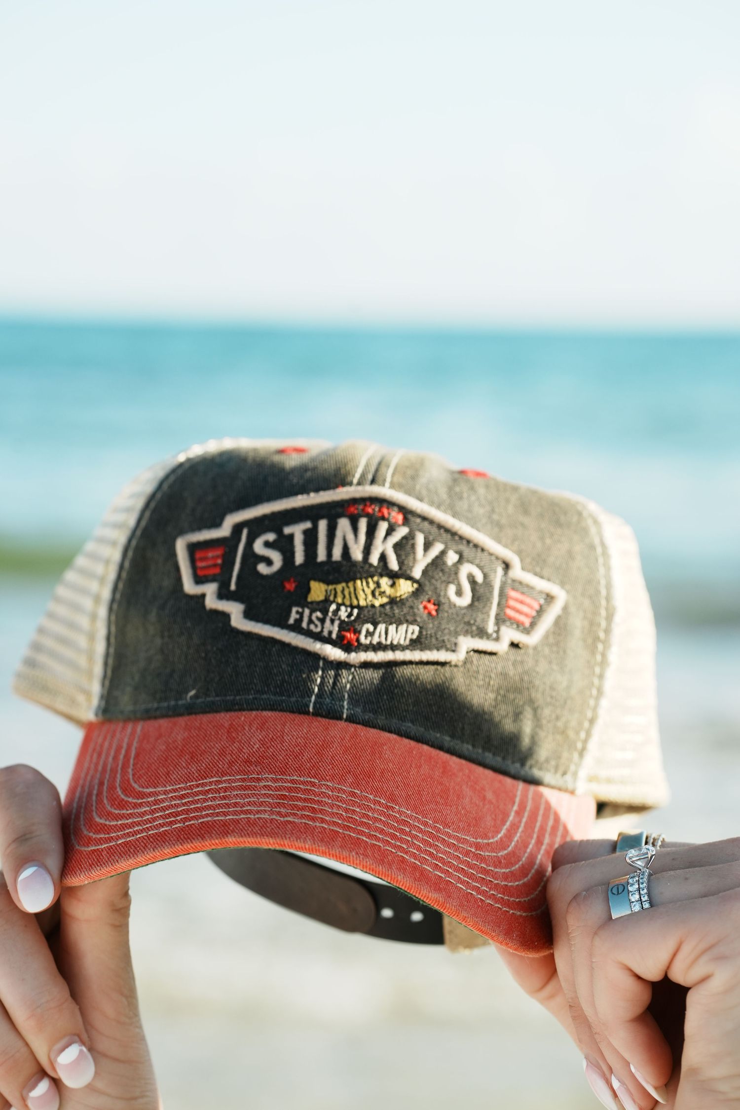 Distressed trucker hat with Stinky's Fish Camp logo in olive green and coral red colors against beach backdrop.