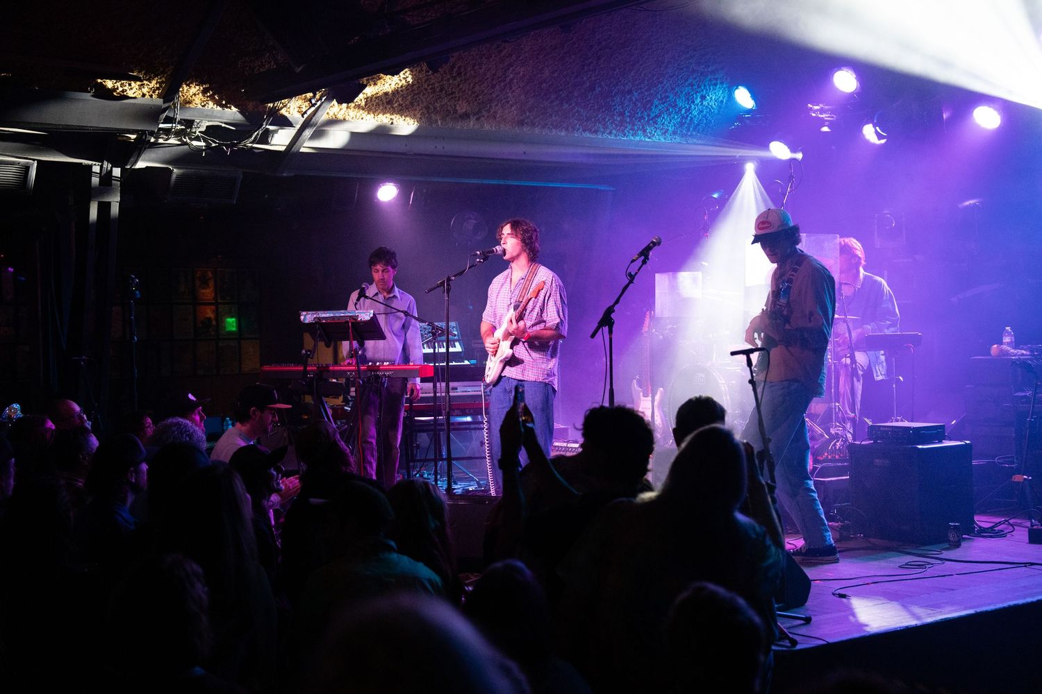Musicians perform on stage with colorful purple and blue lighting effects during a live concert performance.
